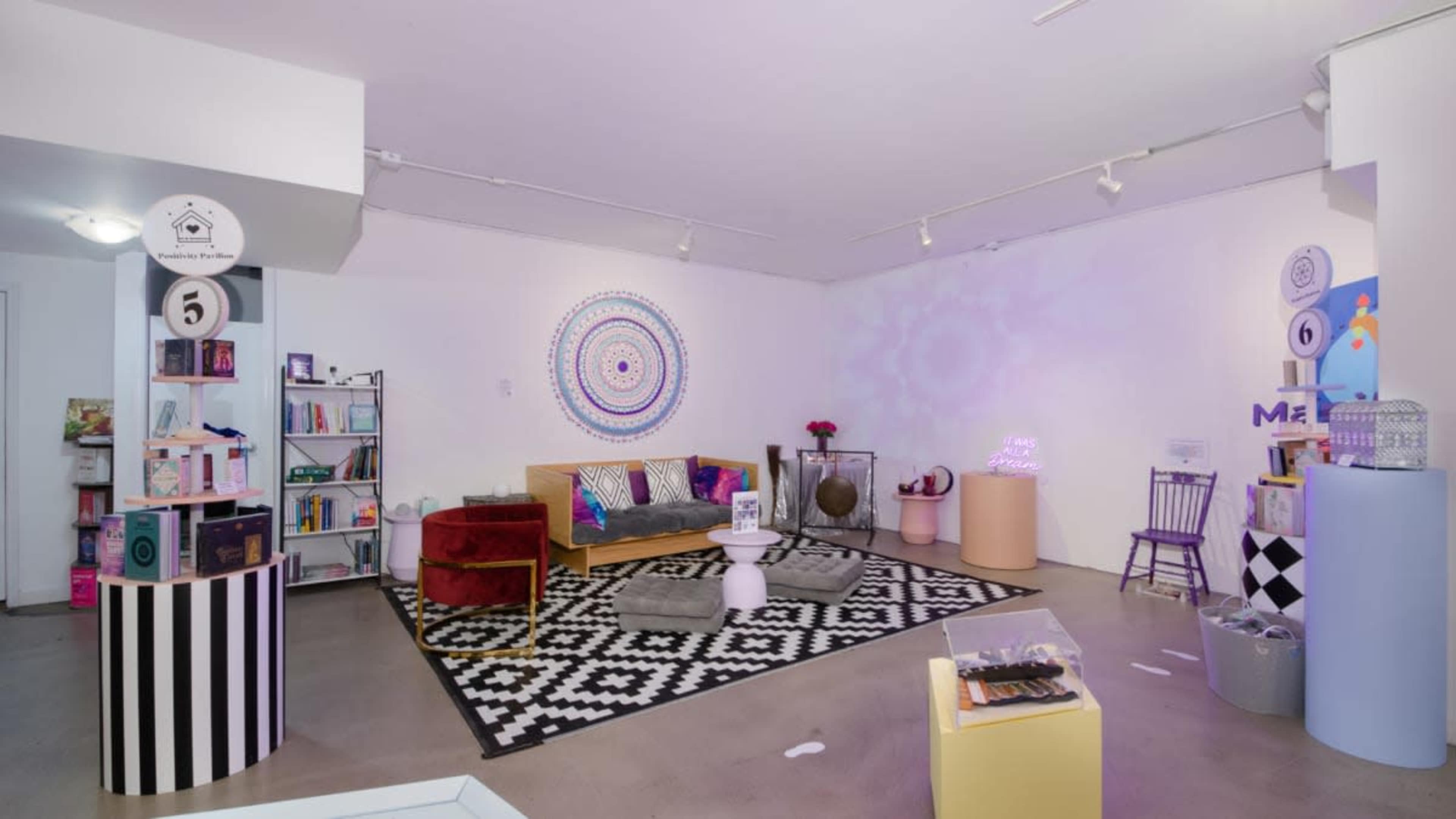 The image shows a modern, brightly lit interior space featuring a colorful rug, a couch, decorative shelves with books, and various artistic displays.