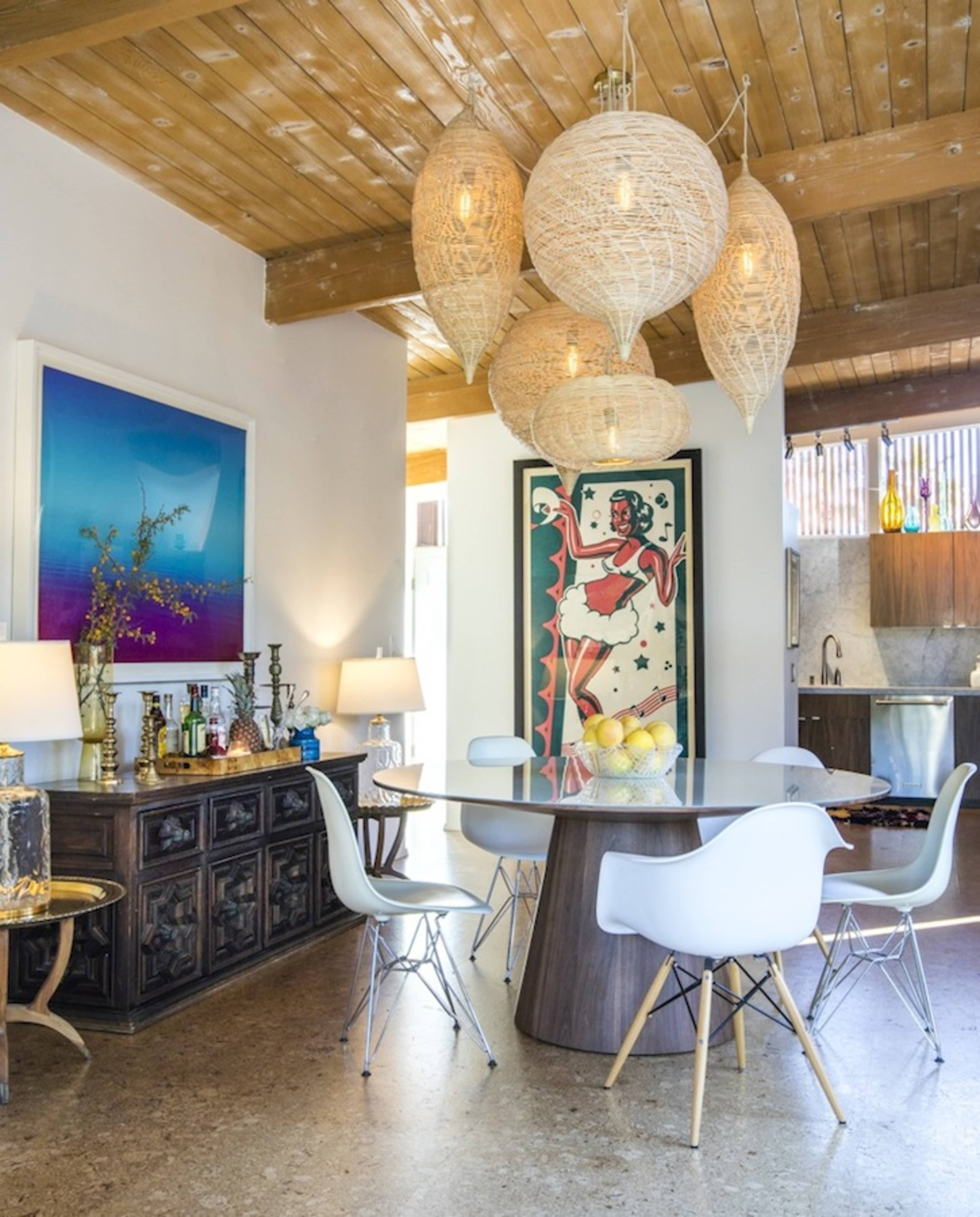A dining area features a round table surrounded by white chairs, illuminated by large woven pendant lights, with a decorative sideboard and colorful artwork on the walls.