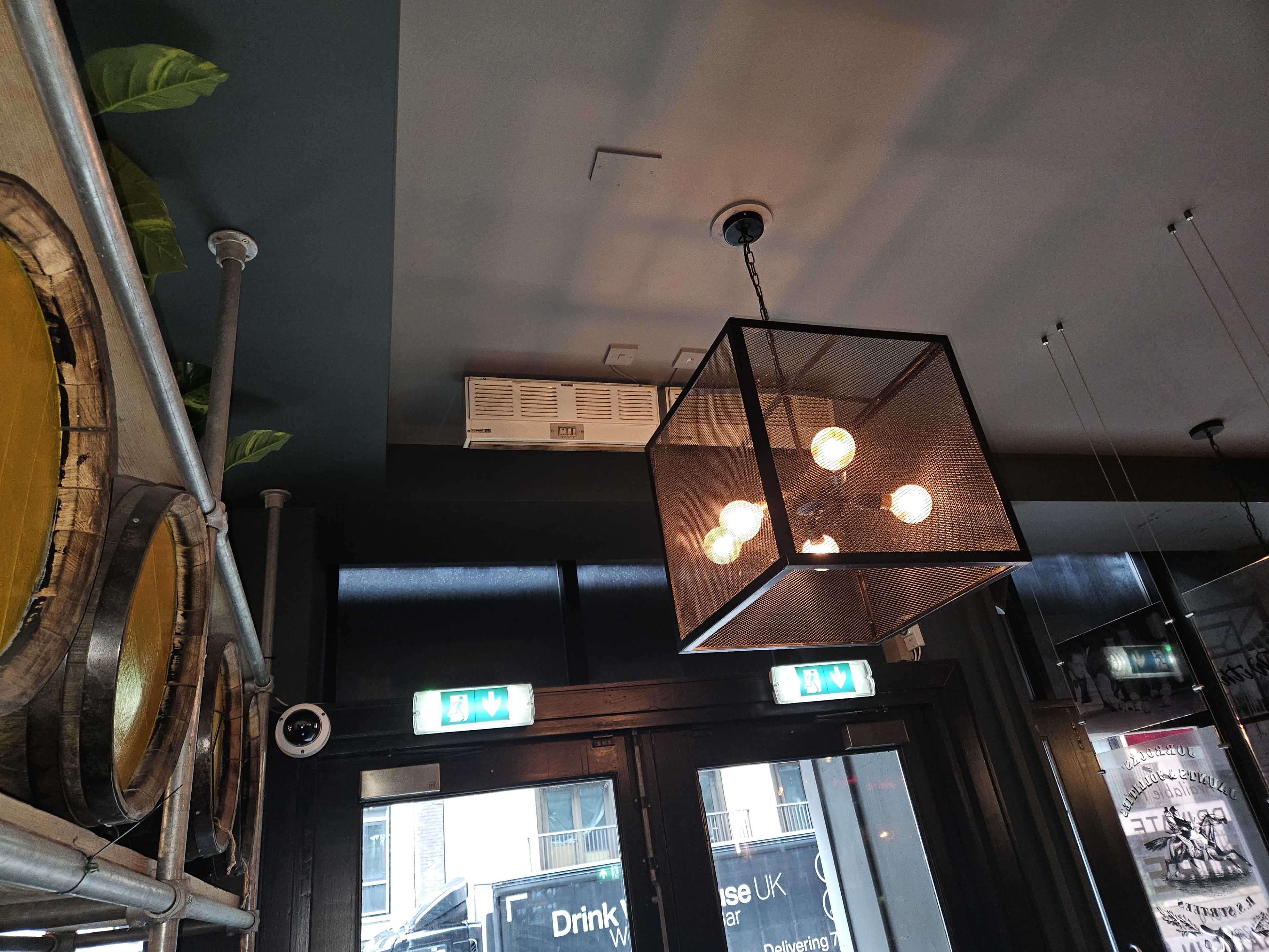 The interior of a bar or restaurant features a large, industrial-style light fixture hanging from the ceiling, with visible air conditioning vents and green plants above.
