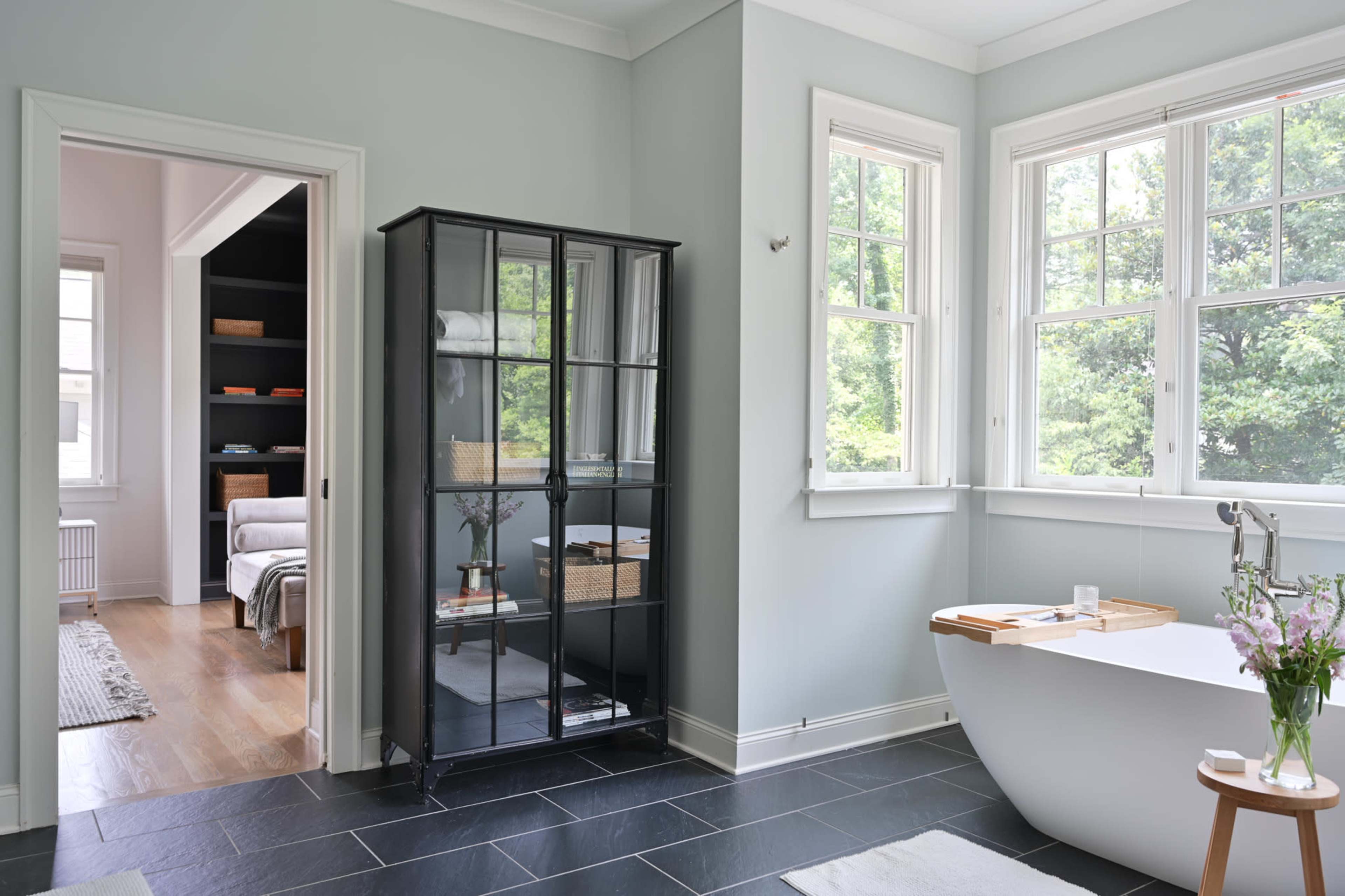 A modern bathroom features a freestanding bathtub, a glass cabinet, and large windows that allow natural light to fill the space.