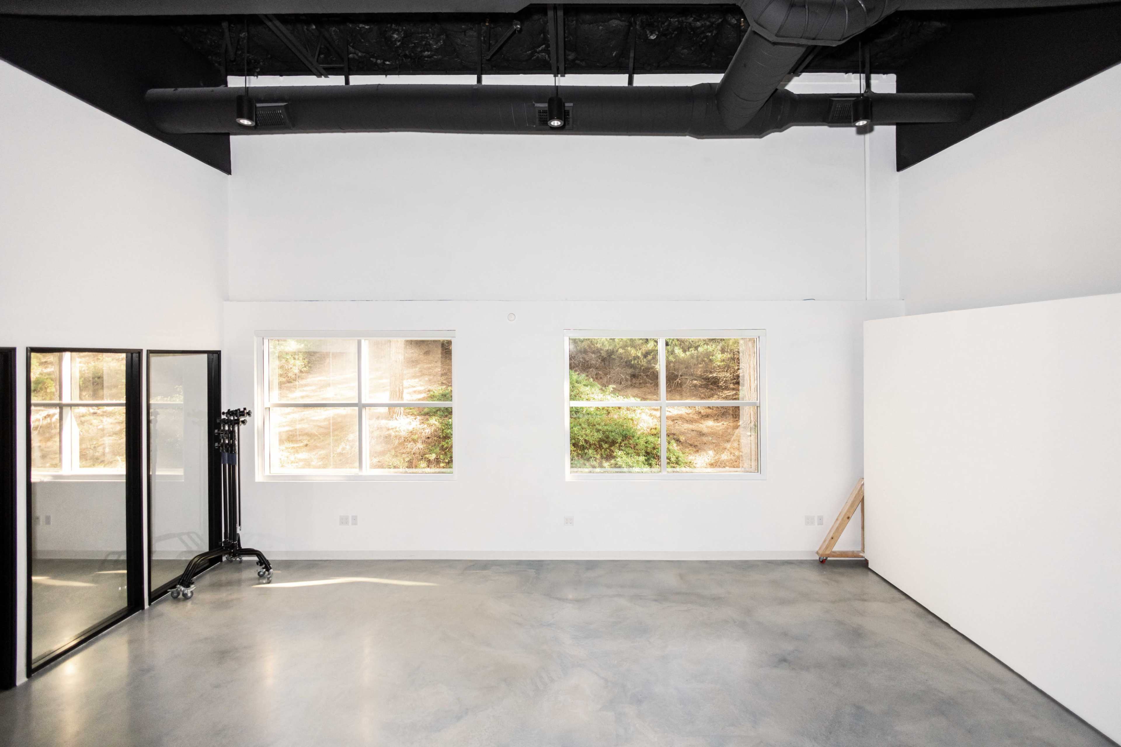 The image shows a spacious, minimalist room with two large windows, a black pipe running along the ceiling, and a mirrored, glass-paneled wall.
