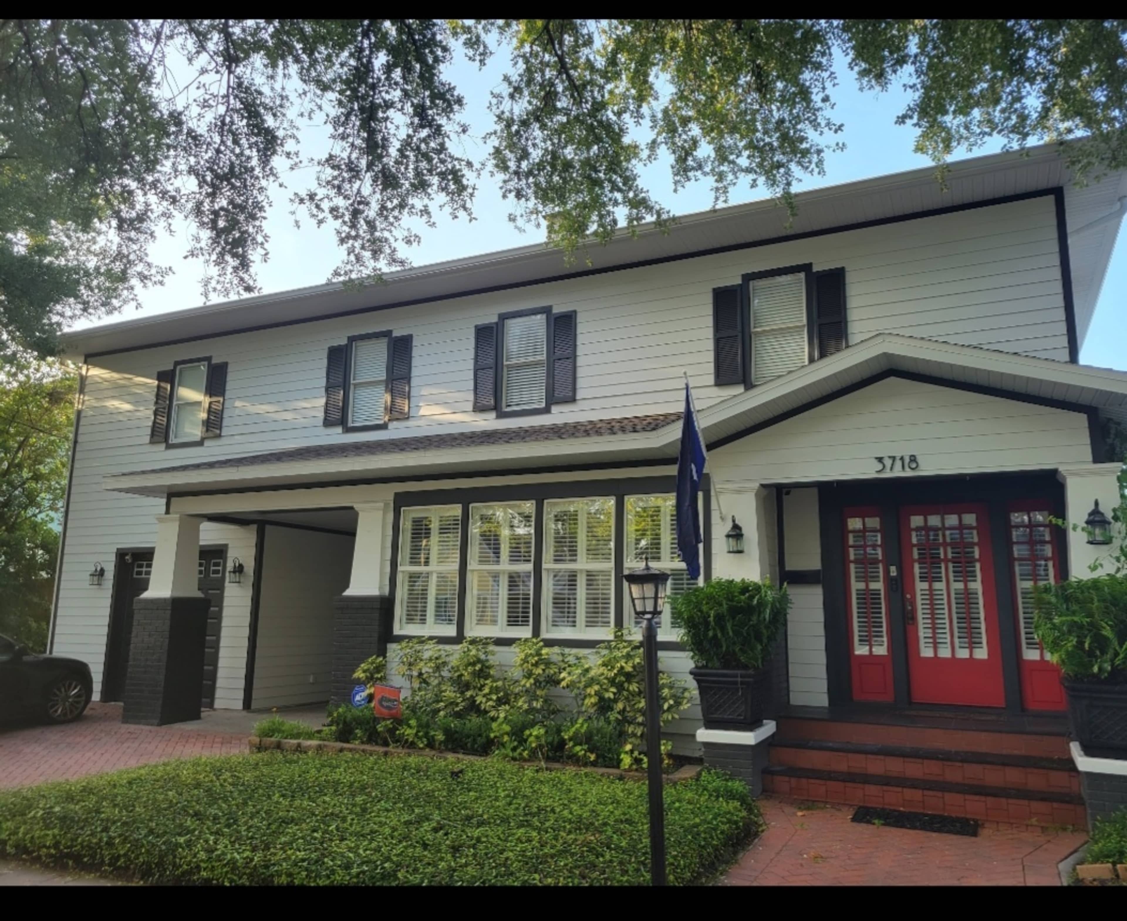 The image shows a two-story house with a white exterior, black shutters, and red double doors, surrounded by landscaping and a brick pathway.
