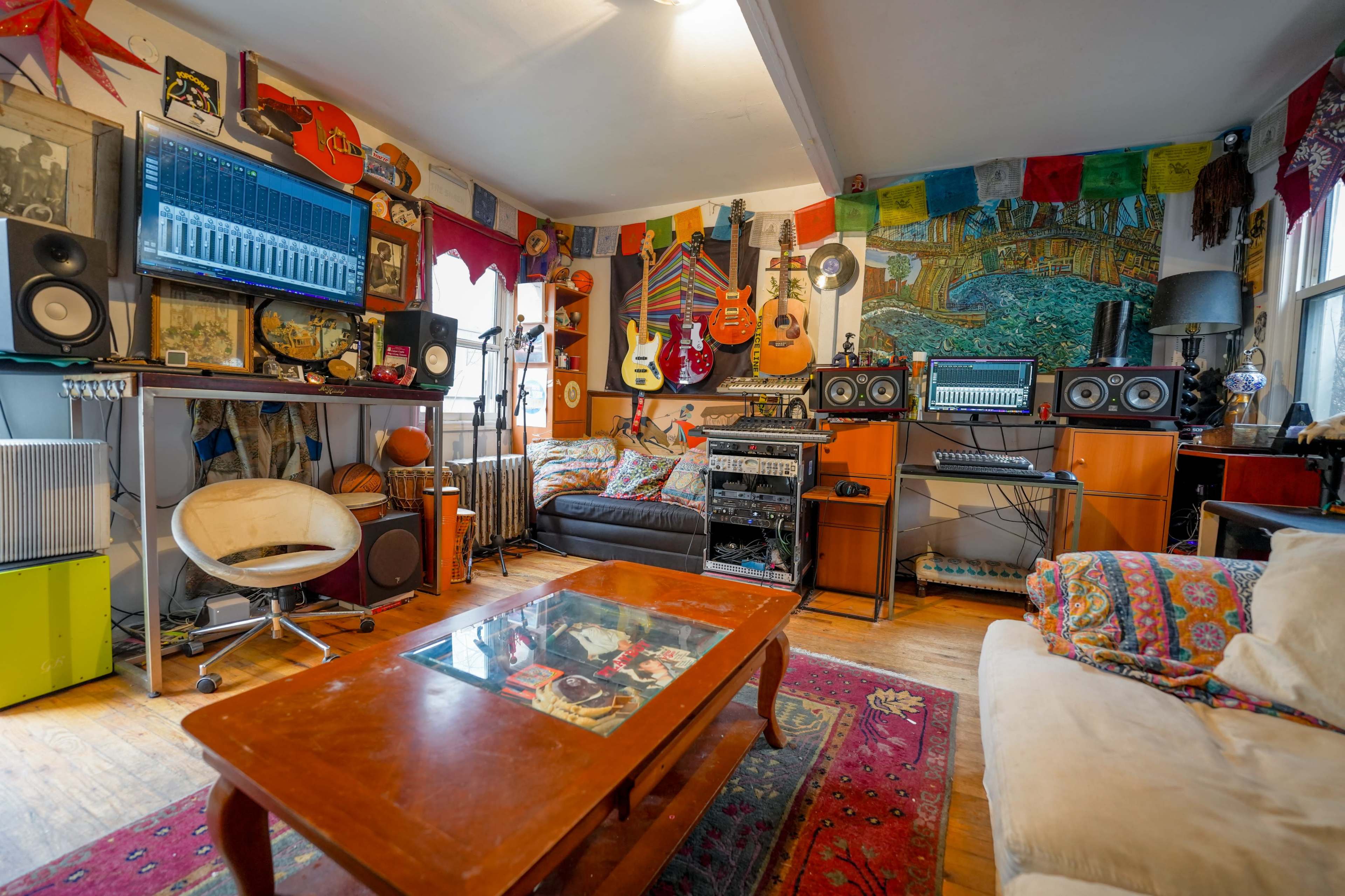 The image shows a colorful music studio filled with guitars, audio equipment, and various decorations, including artwork and textiles.