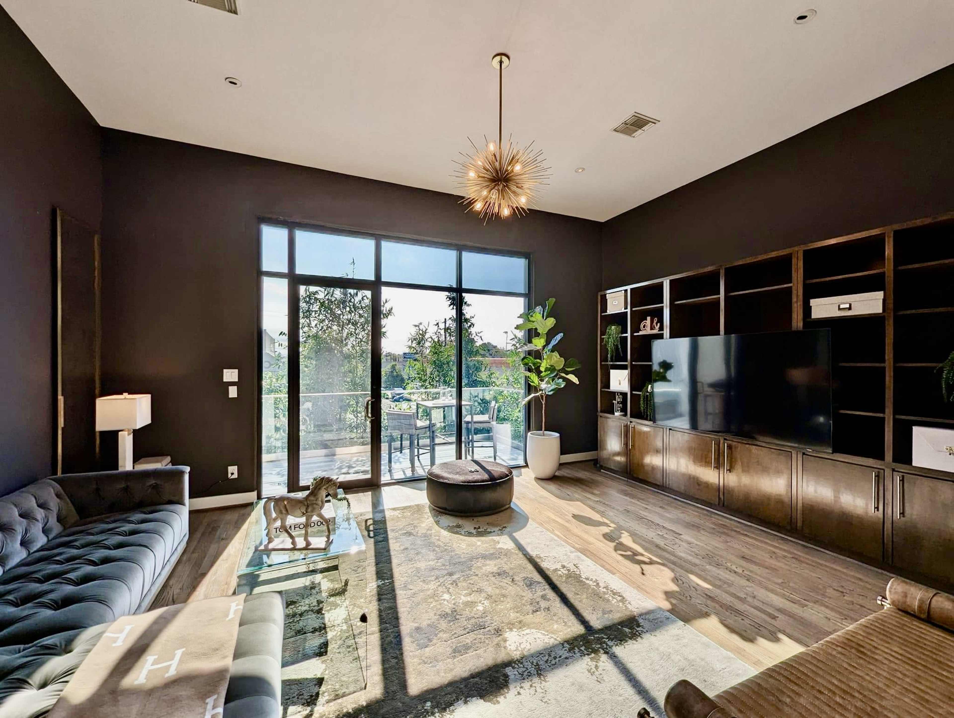 The image shows a modern living room with a large window, a sectional sofa, a round ottoman, and a wall-mounted television.