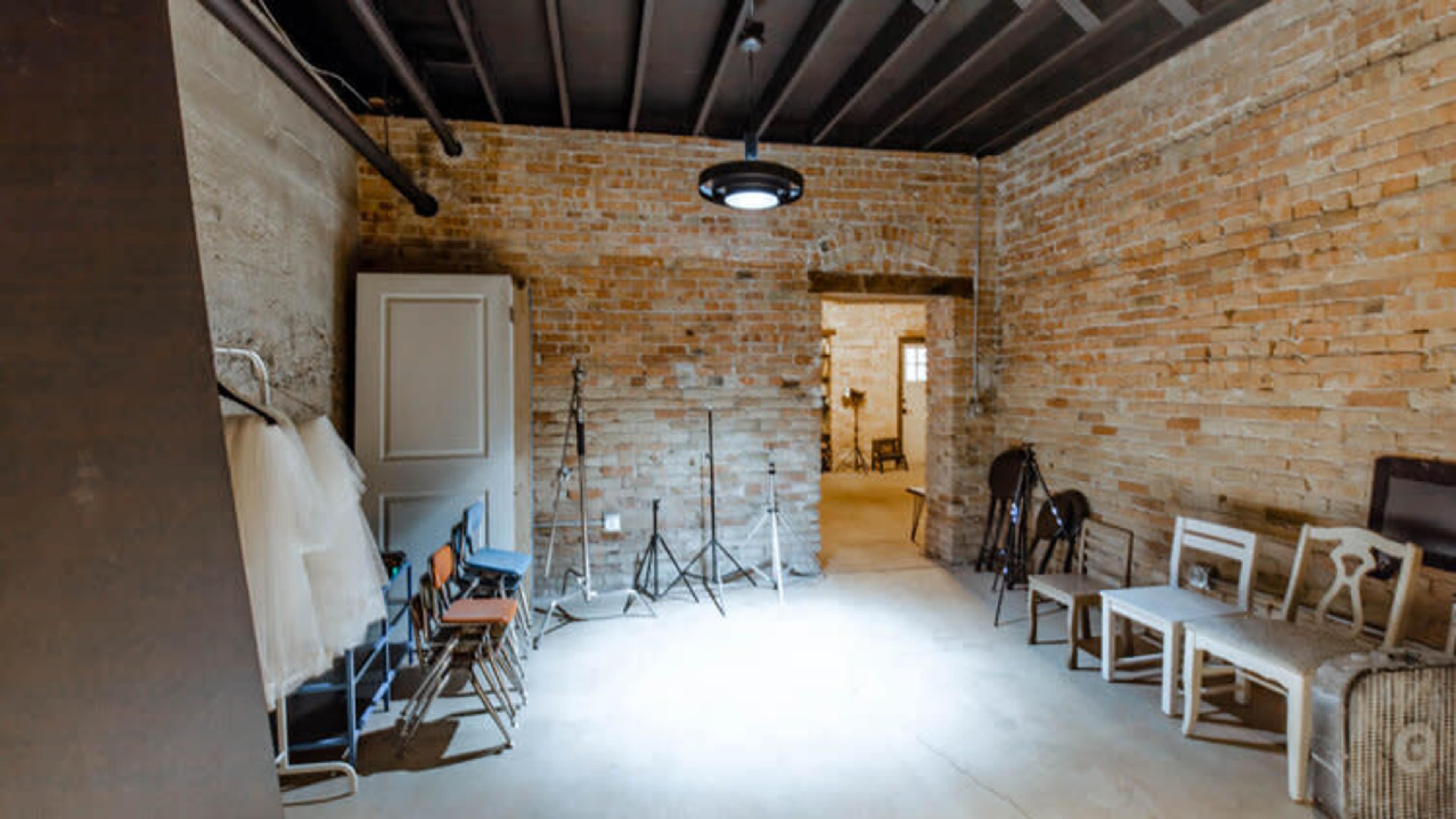 A sparsely furnished room with exposed brick walls, a concrete floor, and several chairs arranged against the walls.