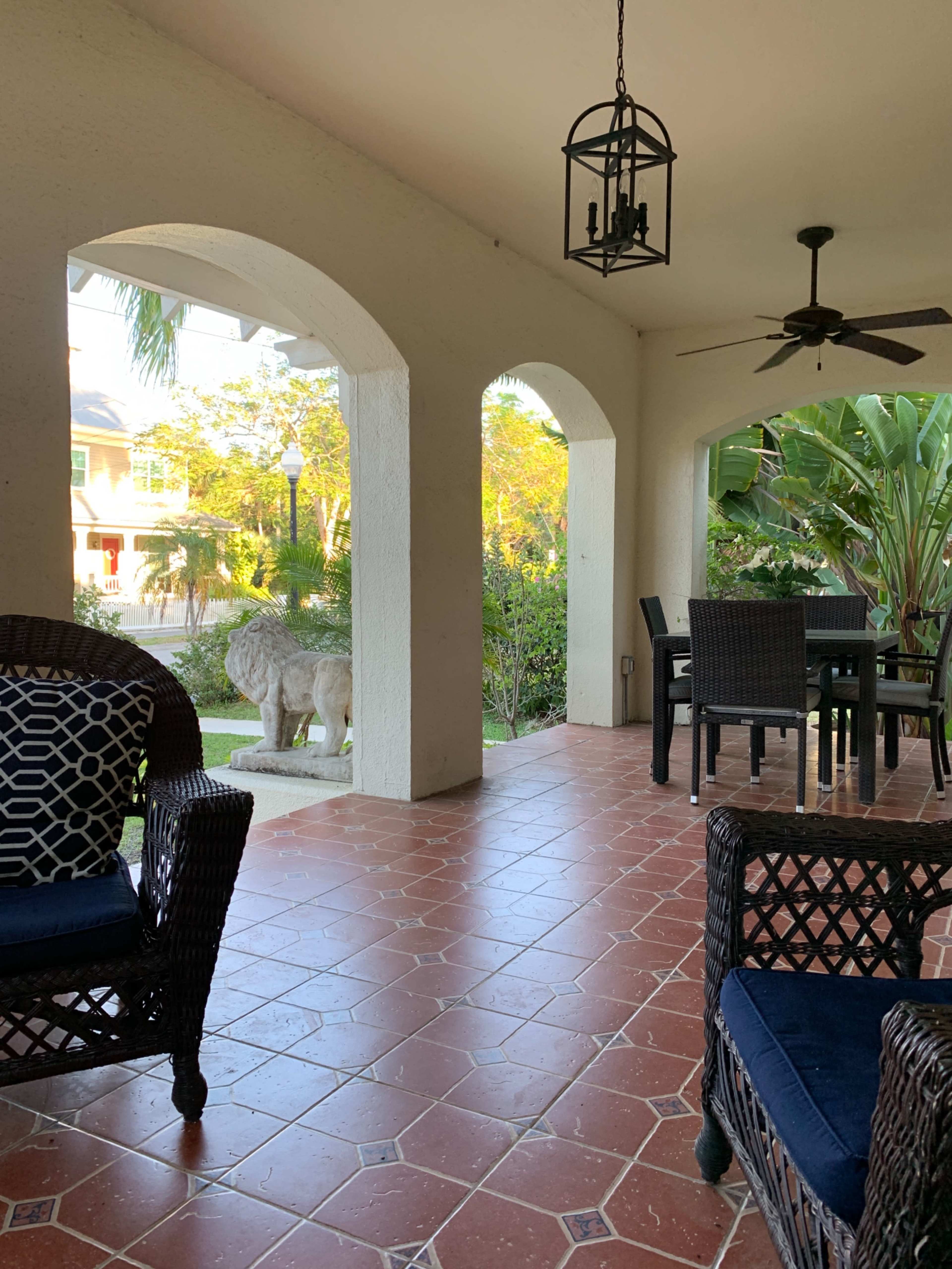 The covered patio features wicker chairs, a table, and a stone lion statue surrounded by tropical plants.