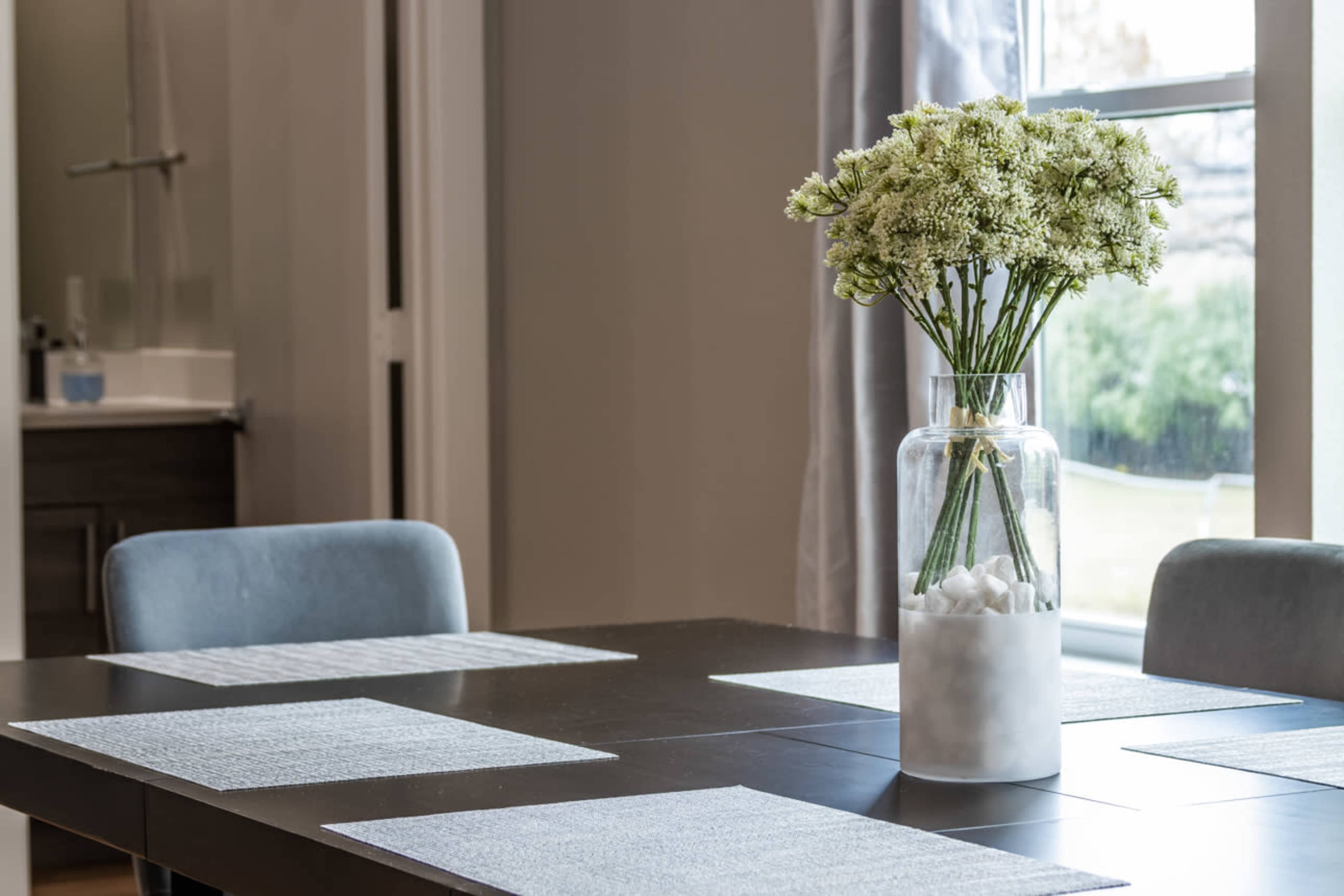 A dining table is set with light-colored placemats, and a glass vase filled with flowers sits at the center.