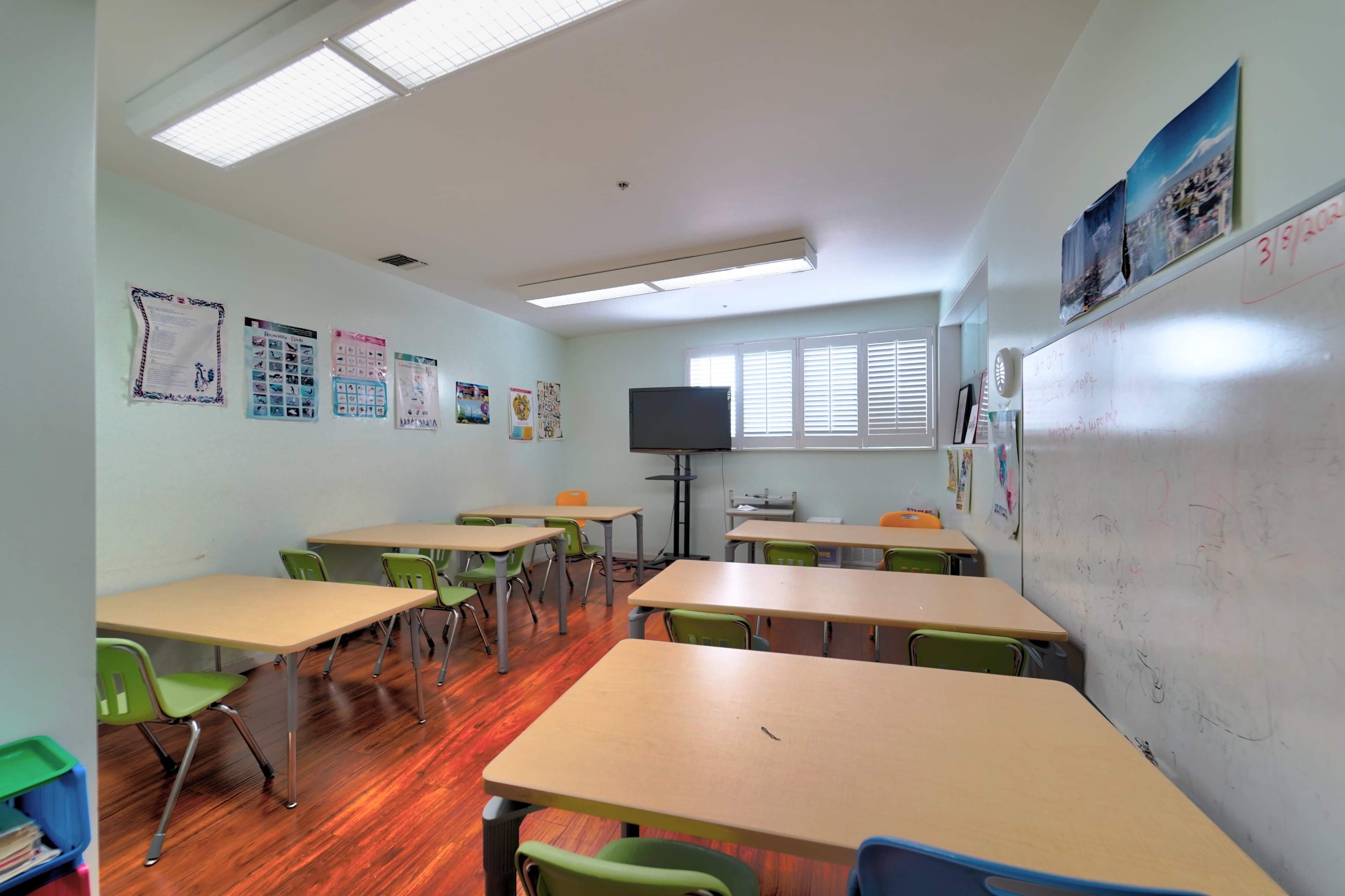 A classroom features several tables with chairs, a large screen, and educational posters on the walls.