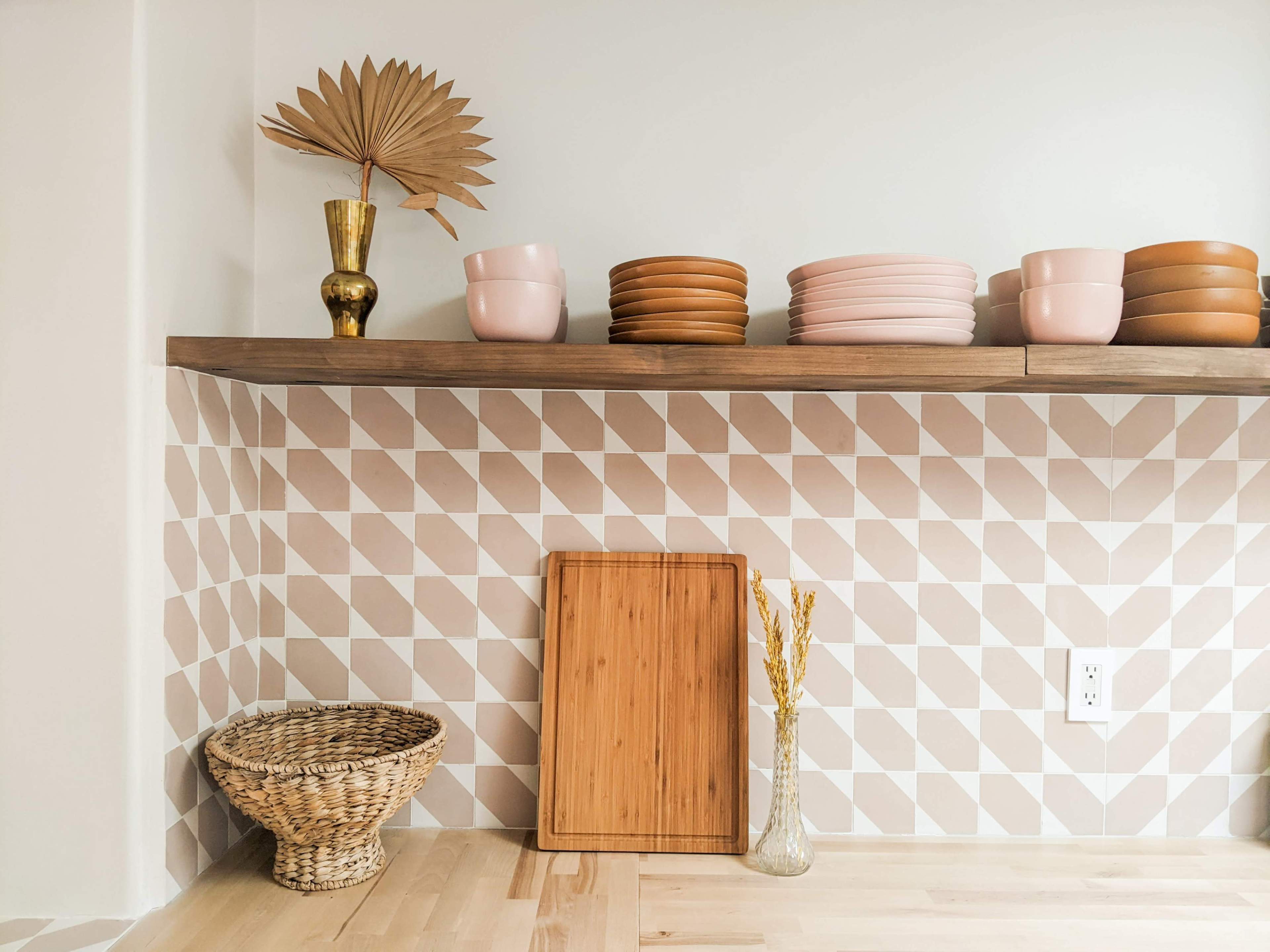 A wooden shelf displays pink plates and bowls above a patterned wall, alongside a woven basket and a bamboo cutting board.