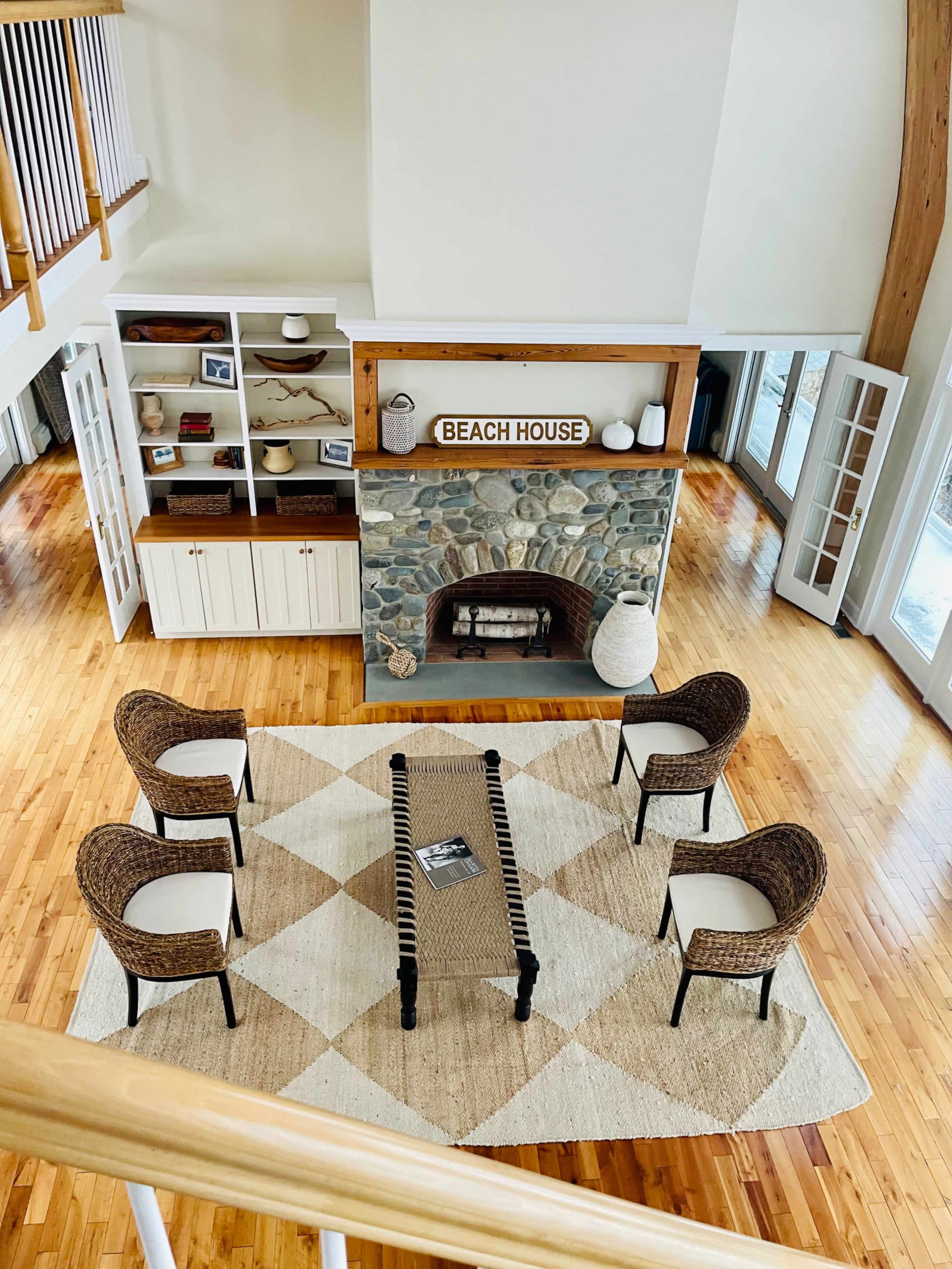 The image shows a spacious room with a stone fireplace, five wicker chairs arranged around a black coffee table on a patterned rug, and large windows letting in natural light.
