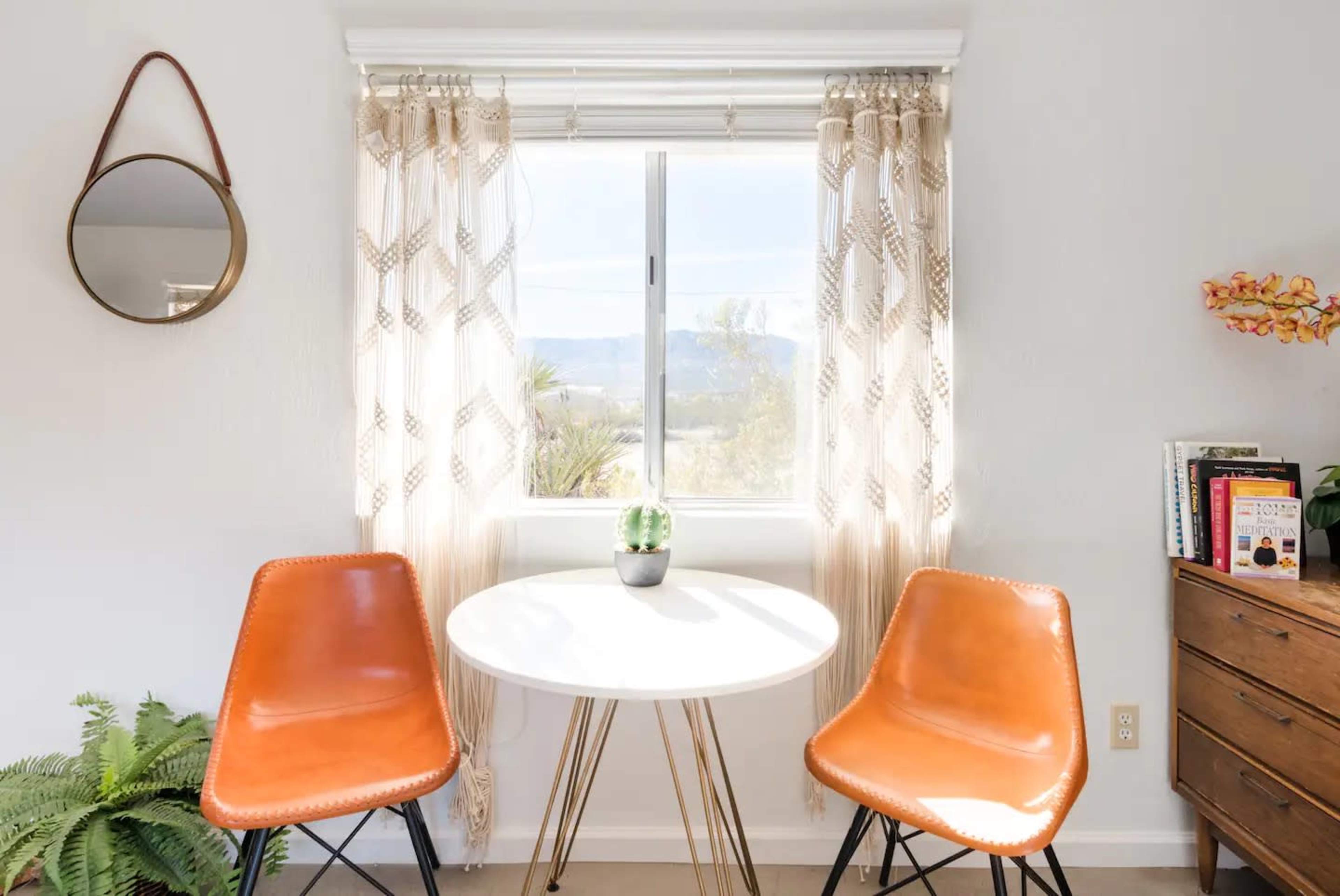 A small dining nook features a round table with two orange chairs, a potted plant on the table, and a window with sheer curtains overlooking a mountainous landscape.