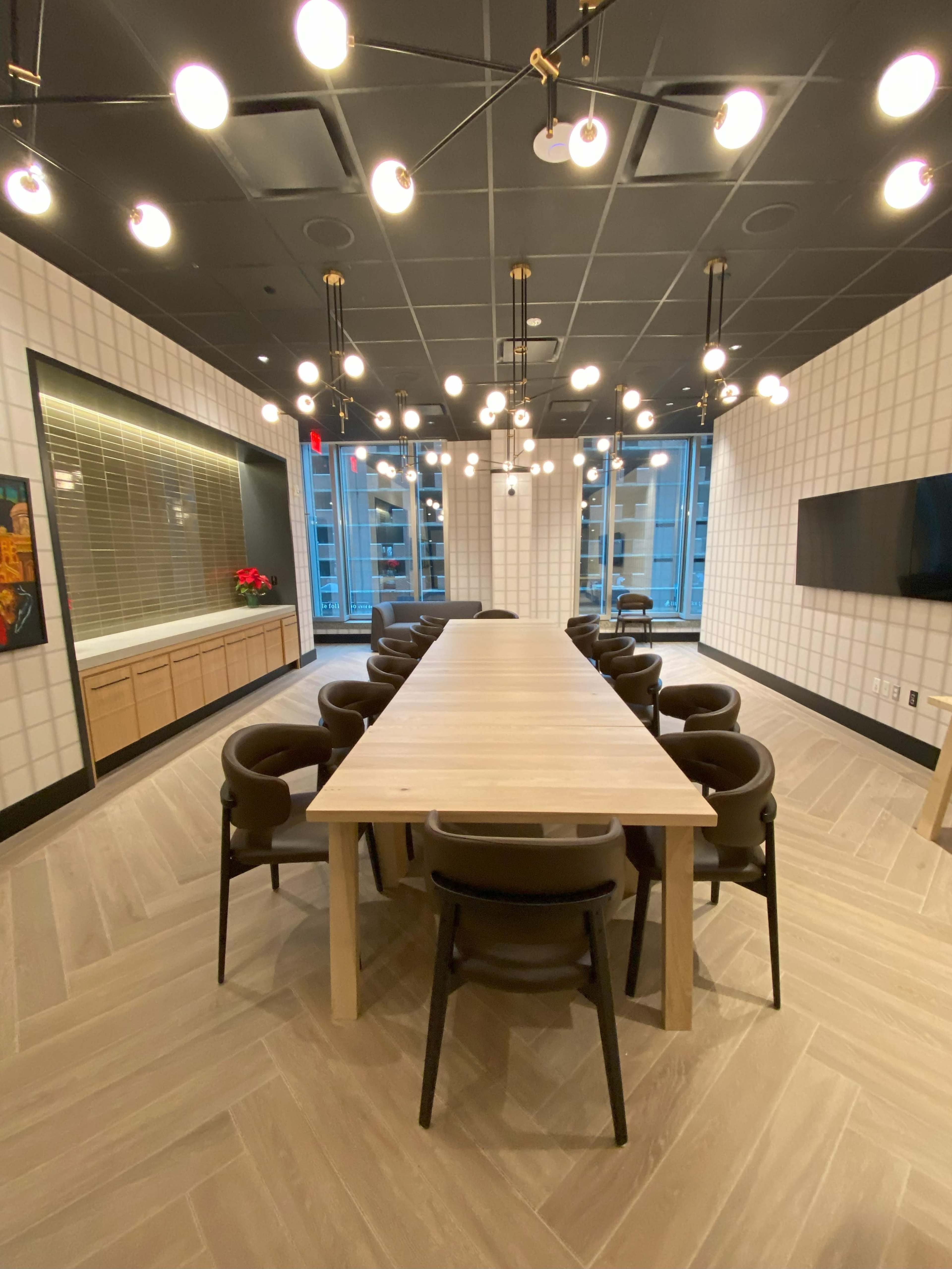 A spacious conference room features a long wooden table surrounded by black chairs, with modern lighting fixtures overhead and large windows in the background.