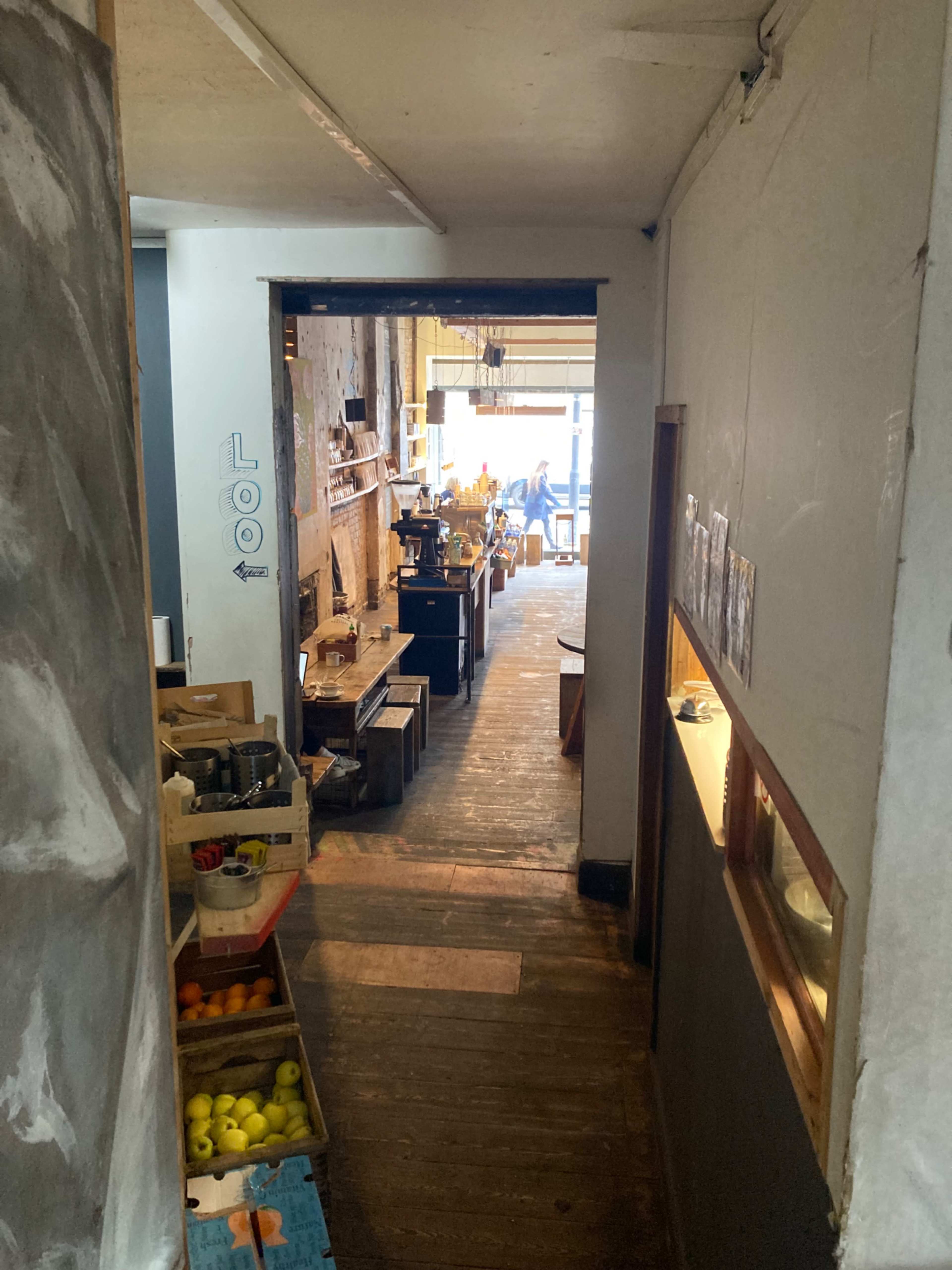 The image shows a hallway leading to a spacious café area with wooden floors and a variety of furnishings.