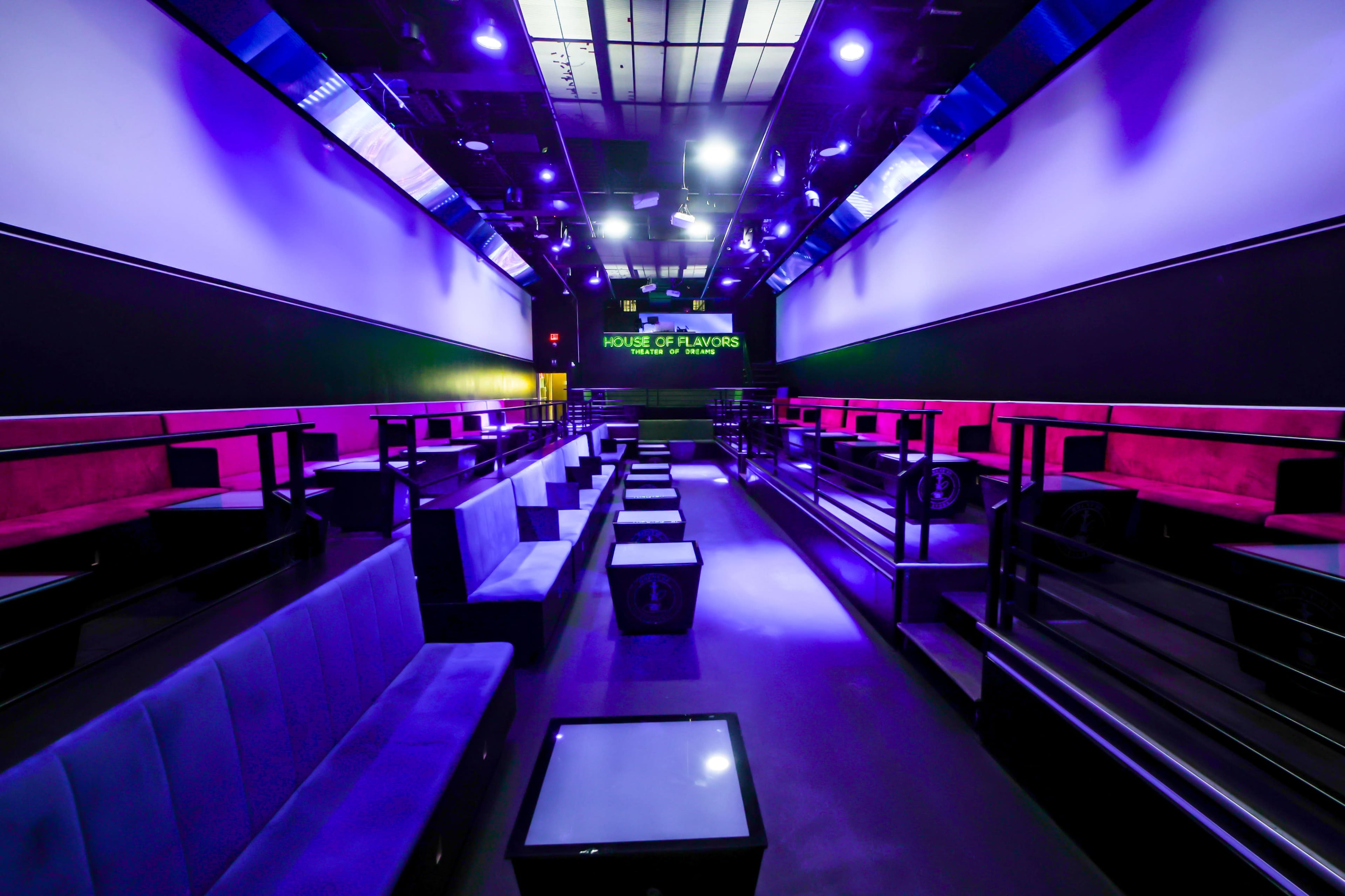 The image shows a modern lounge with dim lighting, featuring rows of seating areas and a large screen displaying "HOUSE OF FLAVORS."