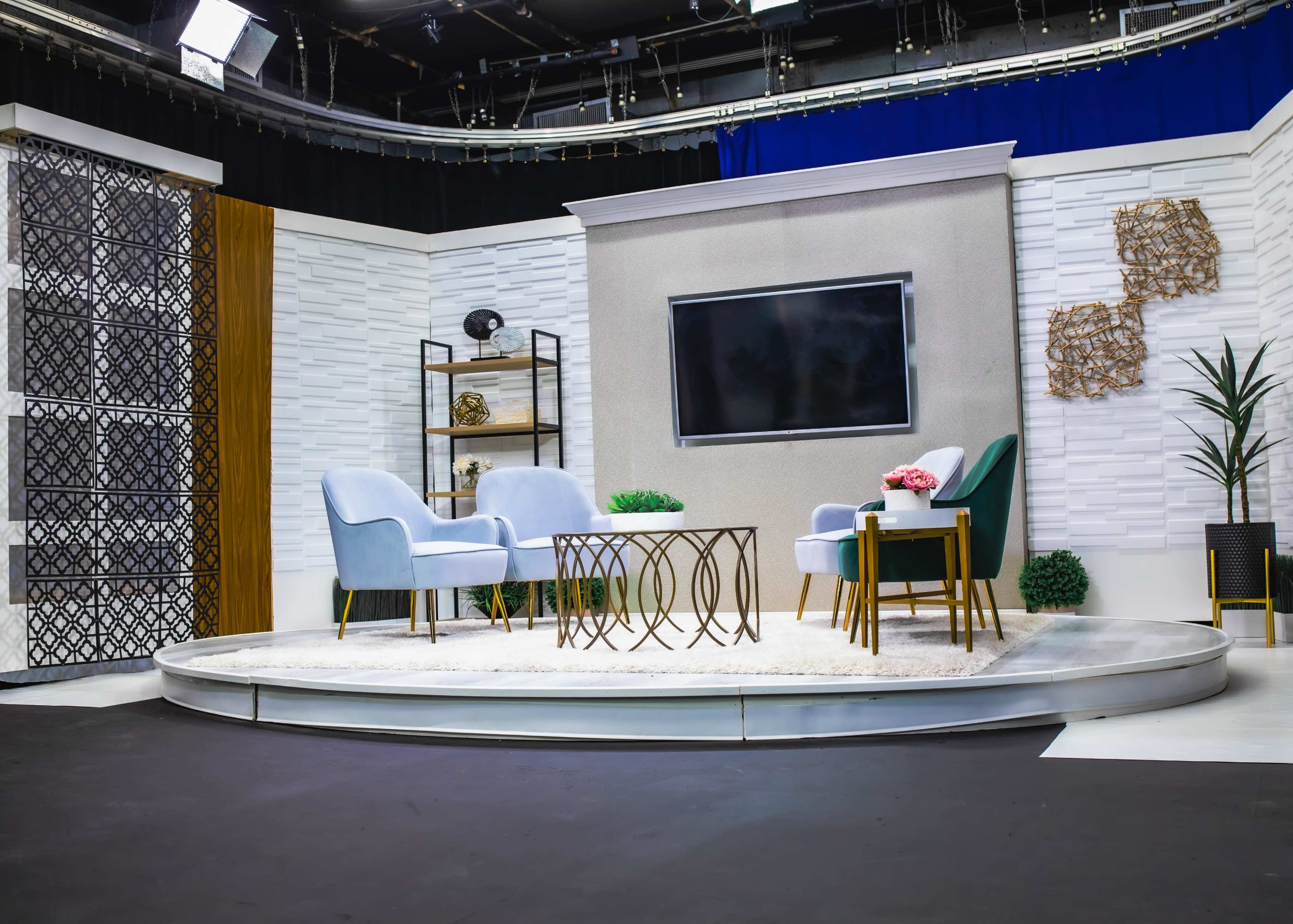 The image shows a modern television studio set with two chairs, a coffee table, and decorative plants arranged on a circular platform.