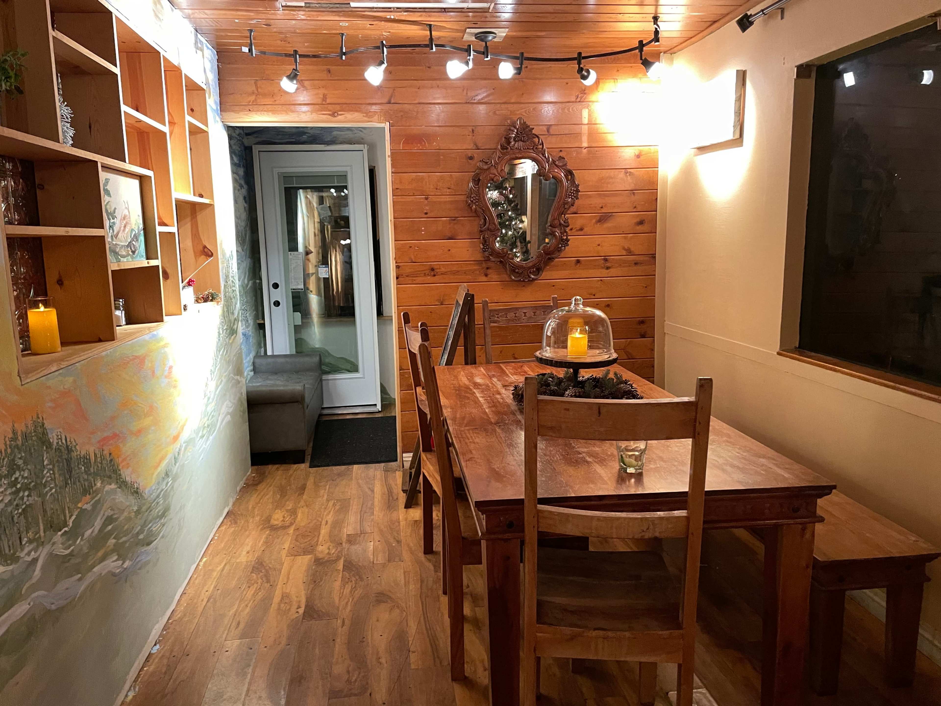 The image shows a wooden dining table with chairs in a warmly lit room that features a large mirror and a door leading to an outdoor area.