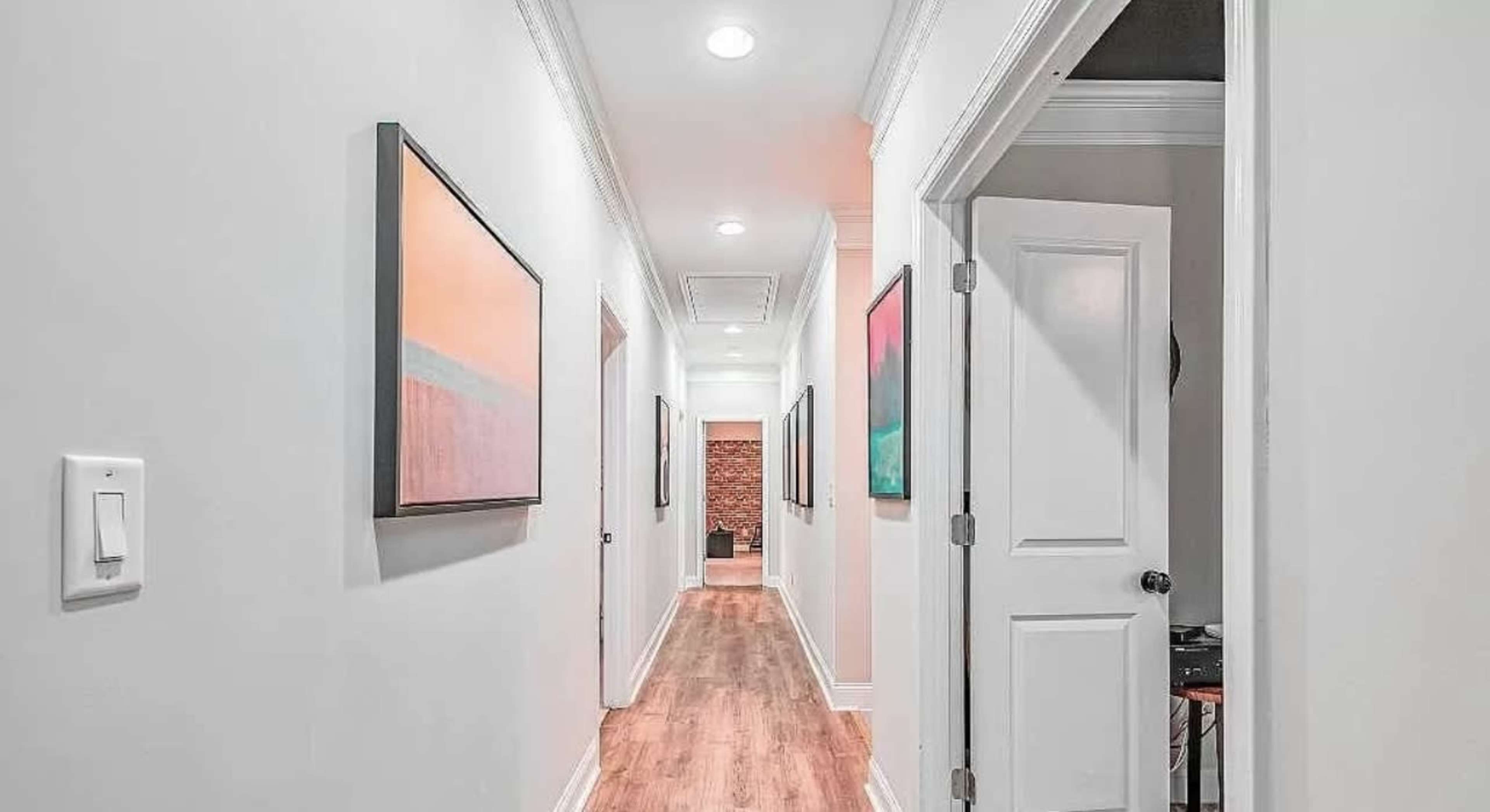 A well-lit hallway with framed artwork on the walls and wooden flooring leads to a doorway at the end.