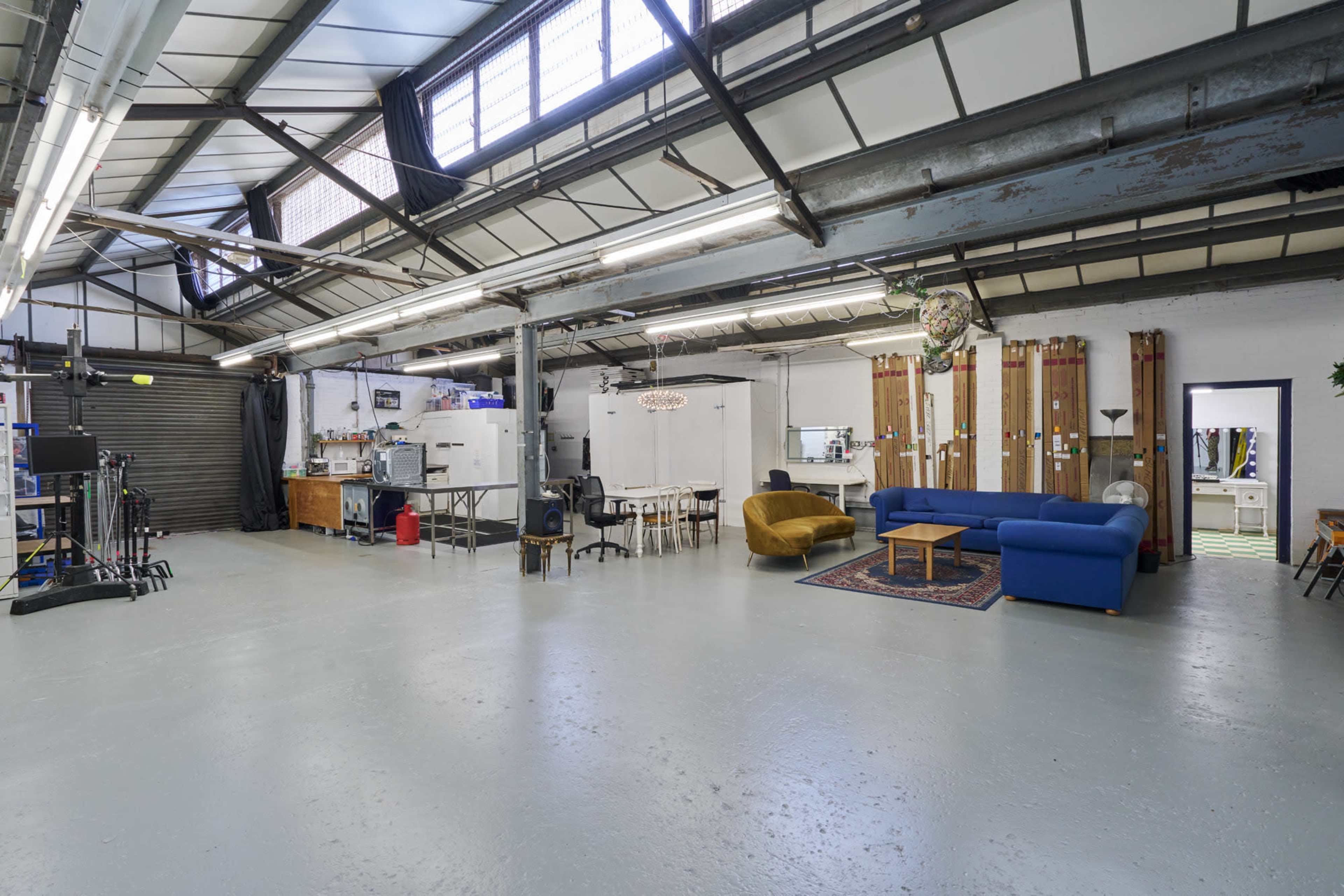 The image shows a spacious industrial-style room with polished concrete flooring, featuring a blue sofa, a wooden table with chairs, and various equipment and furniture along the walls.