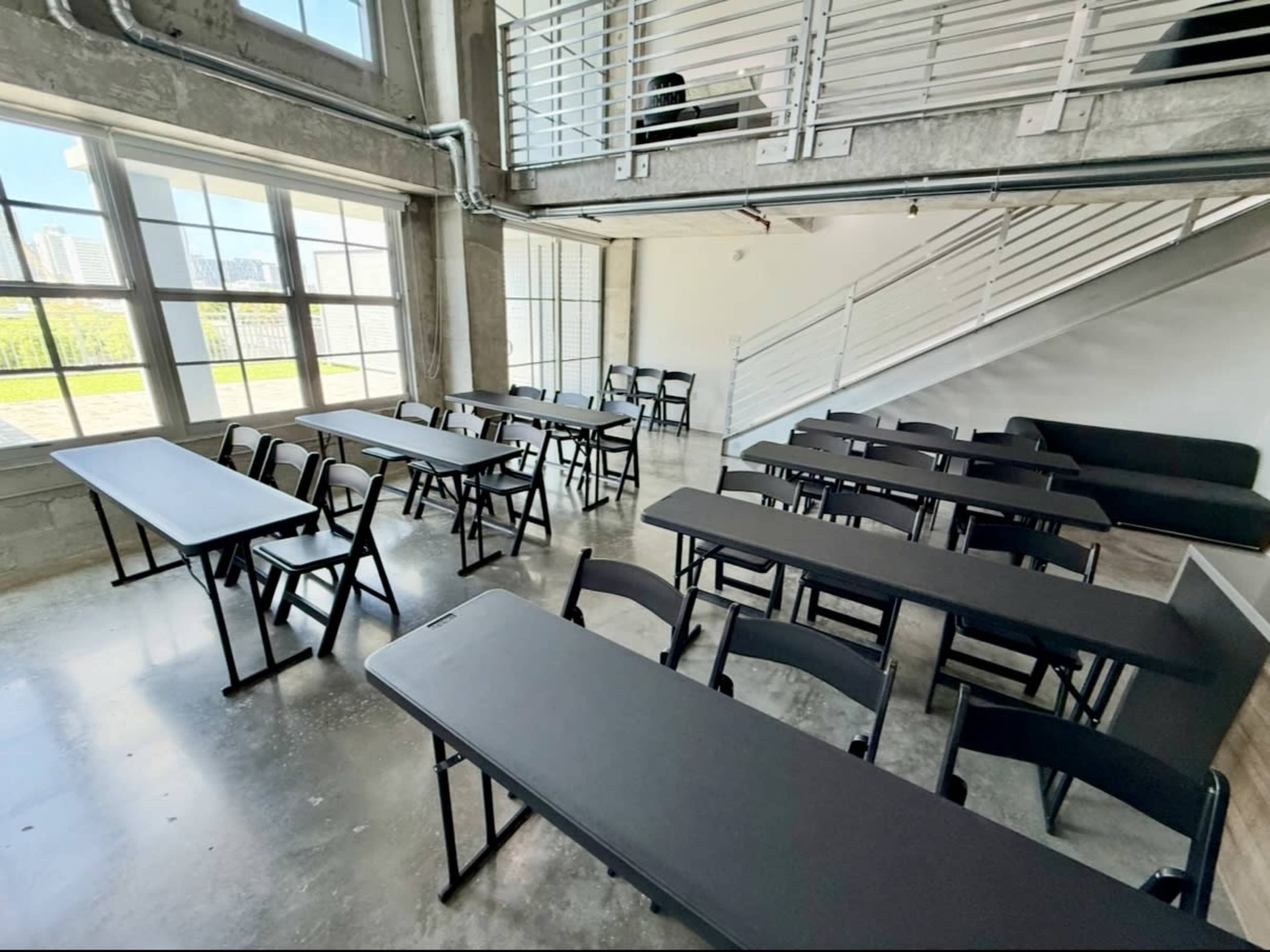 The image shows a modern, airy room with several black folding tables and chairs arranged for a meeting or event, large windows allowing natural light to fill the space.