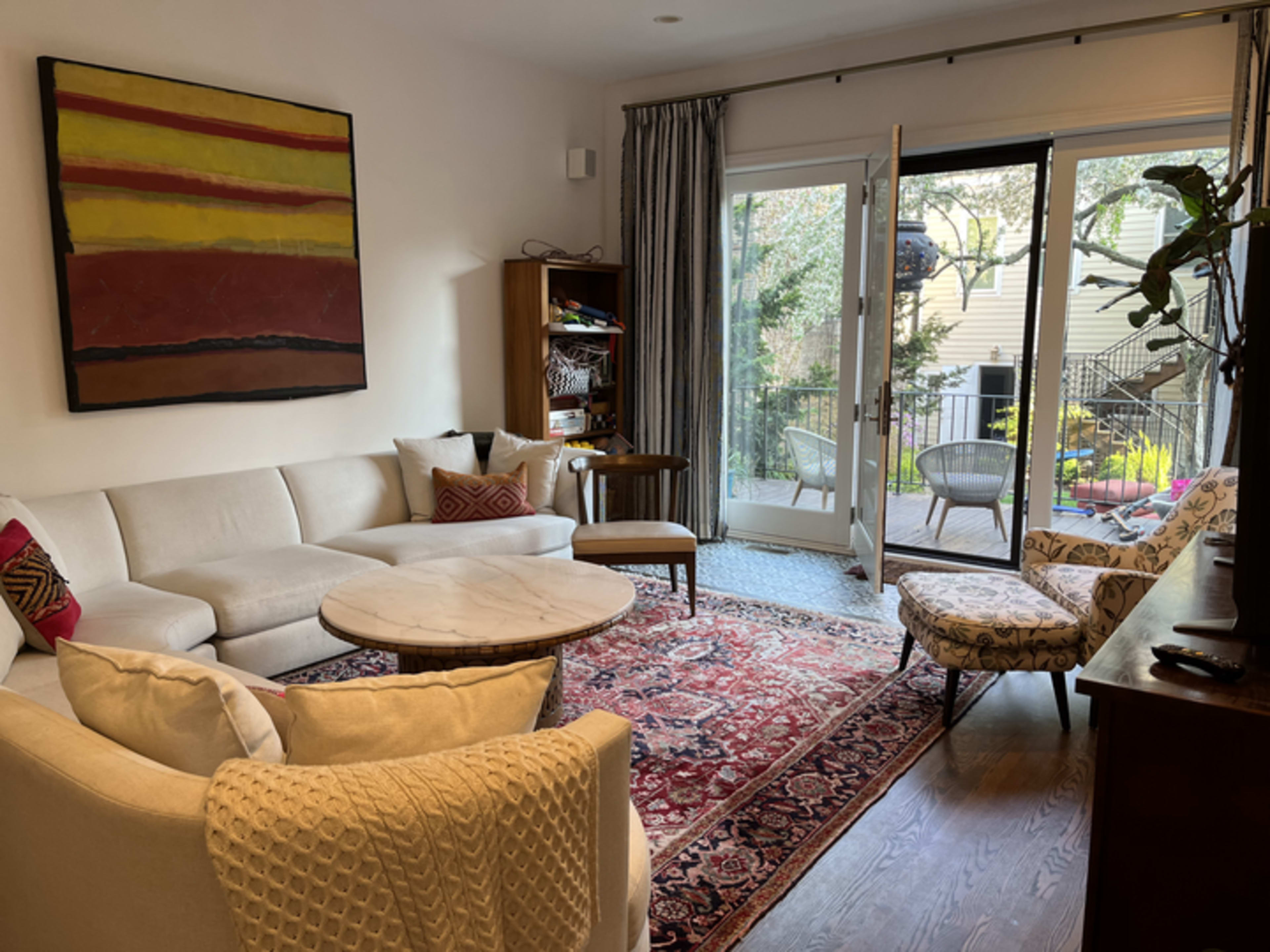 A living room features a sectional sofa, a round coffee table, a patterned rug, and large glass doors leading to a patio area with outdoor seating.