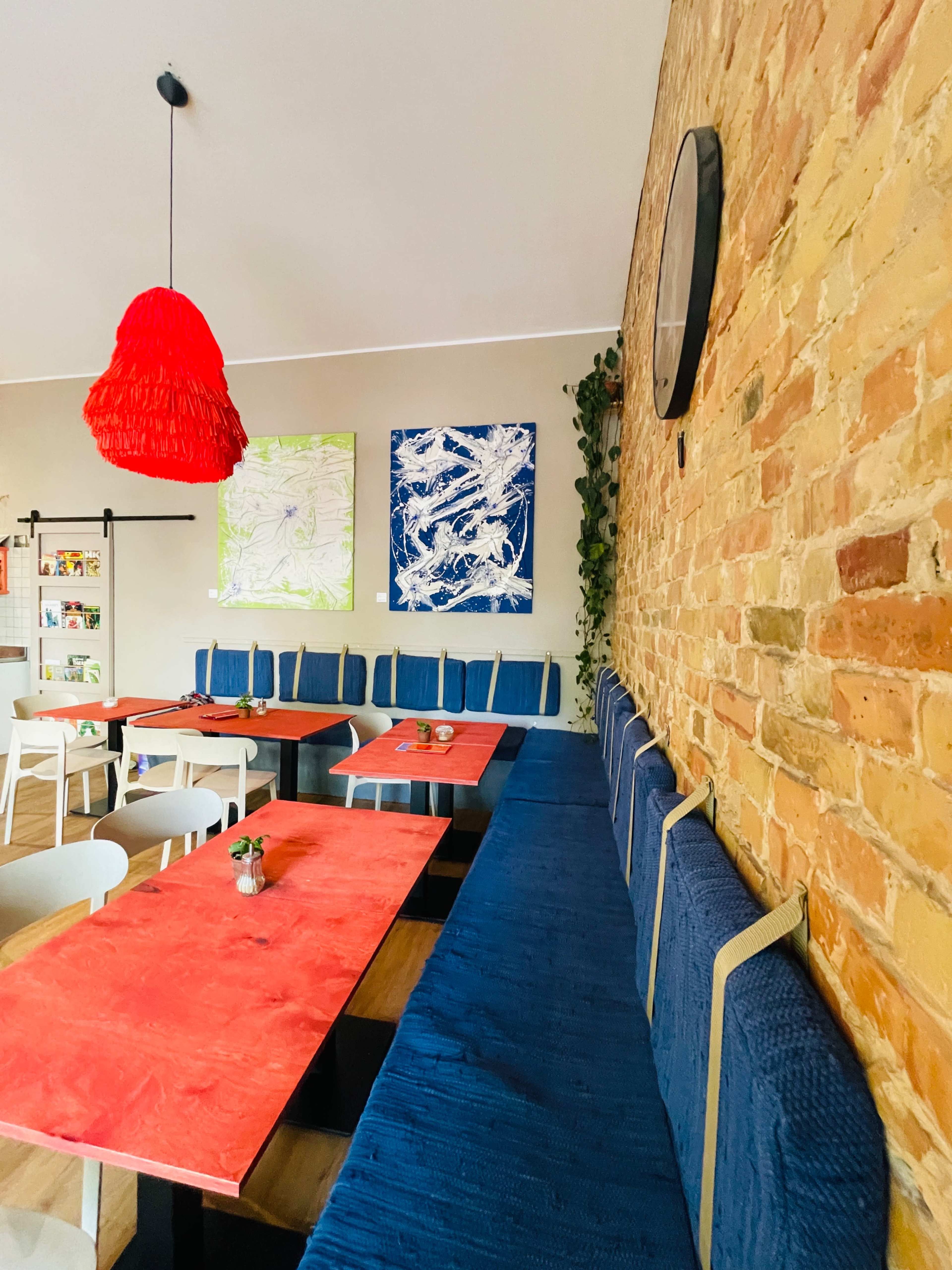The image features a cozy restaurant interior with blue benches and red tabletops, complemented by colorful abstract artworks on the walls.