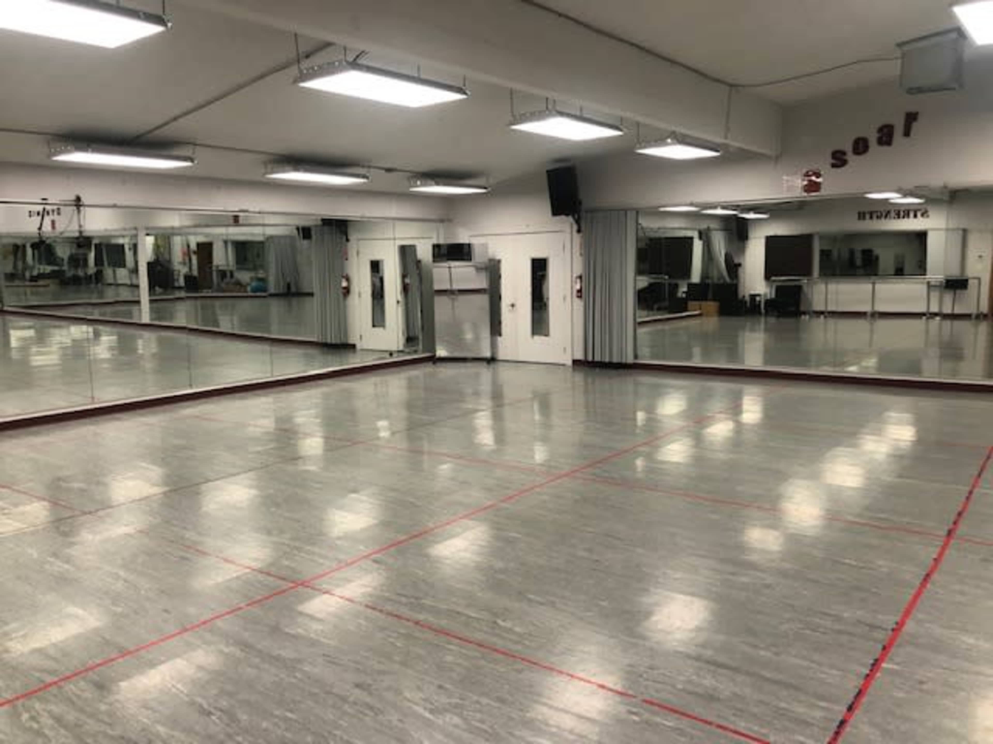 A spacious dance studio featuring mirrored walls, a polished floor, and bright overhead lighting.