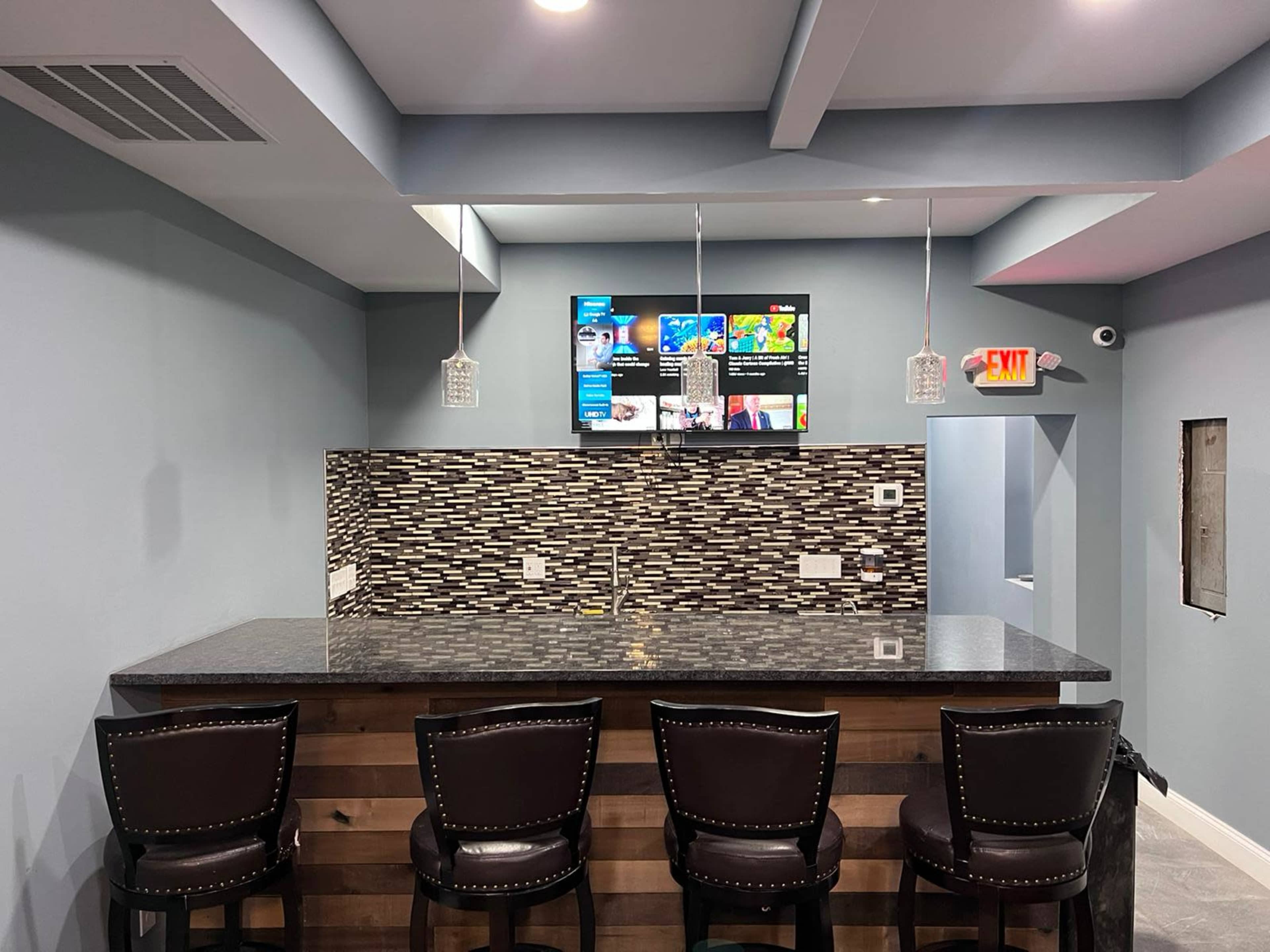 The image shows a modern bar area with a granite countertop, four chairs, and a wall-mounted television displaying various graphics in a room with blue walls.