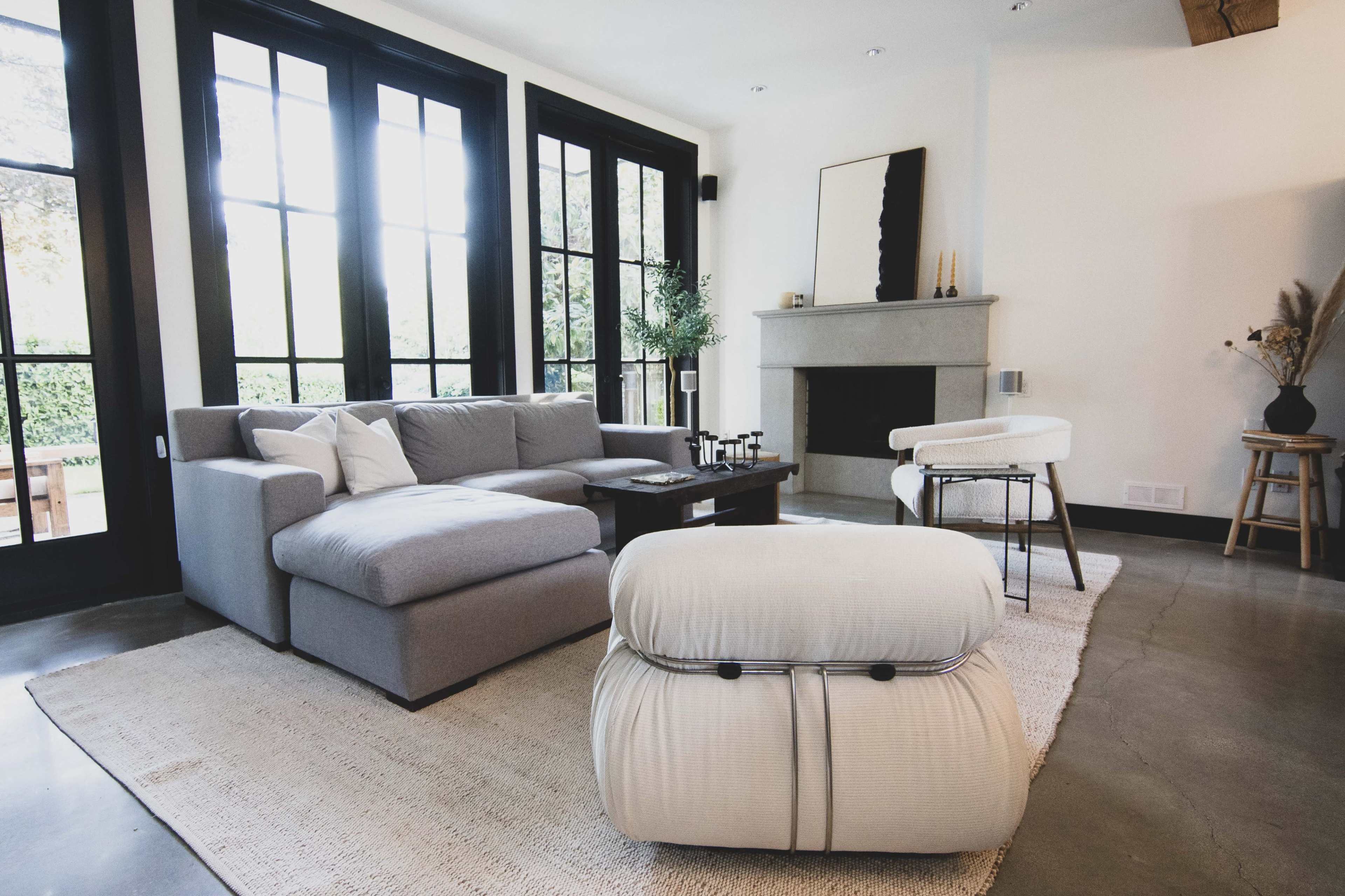 A modern living room with a gray sectional sofa, a wooden coffee table, and large black-framed windows allowing natural light to enter.