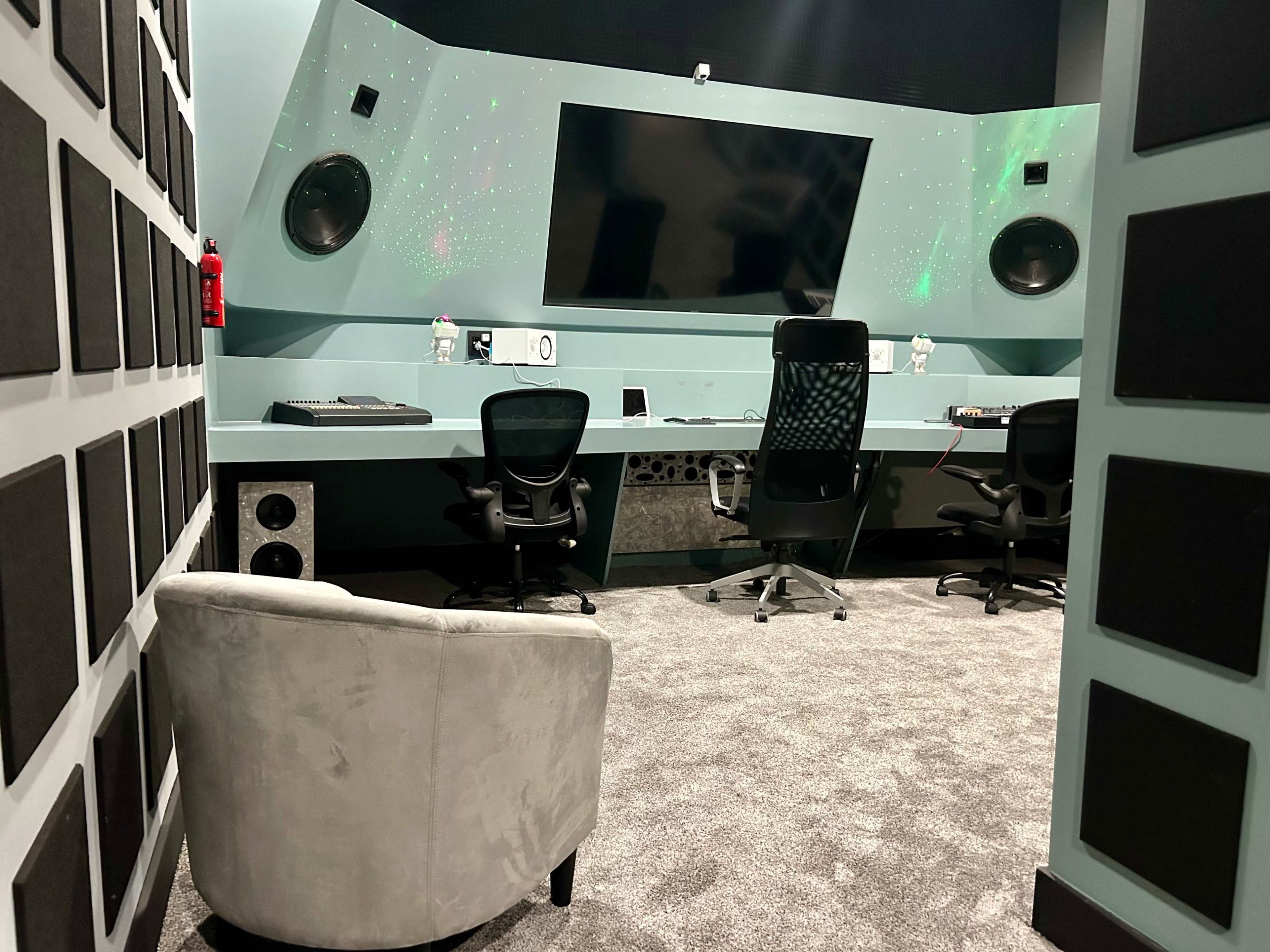 The image shows a sound studio equipped with a large screen, audio equipment, and two office chairs, with a small, plush chair positioned in the corner.