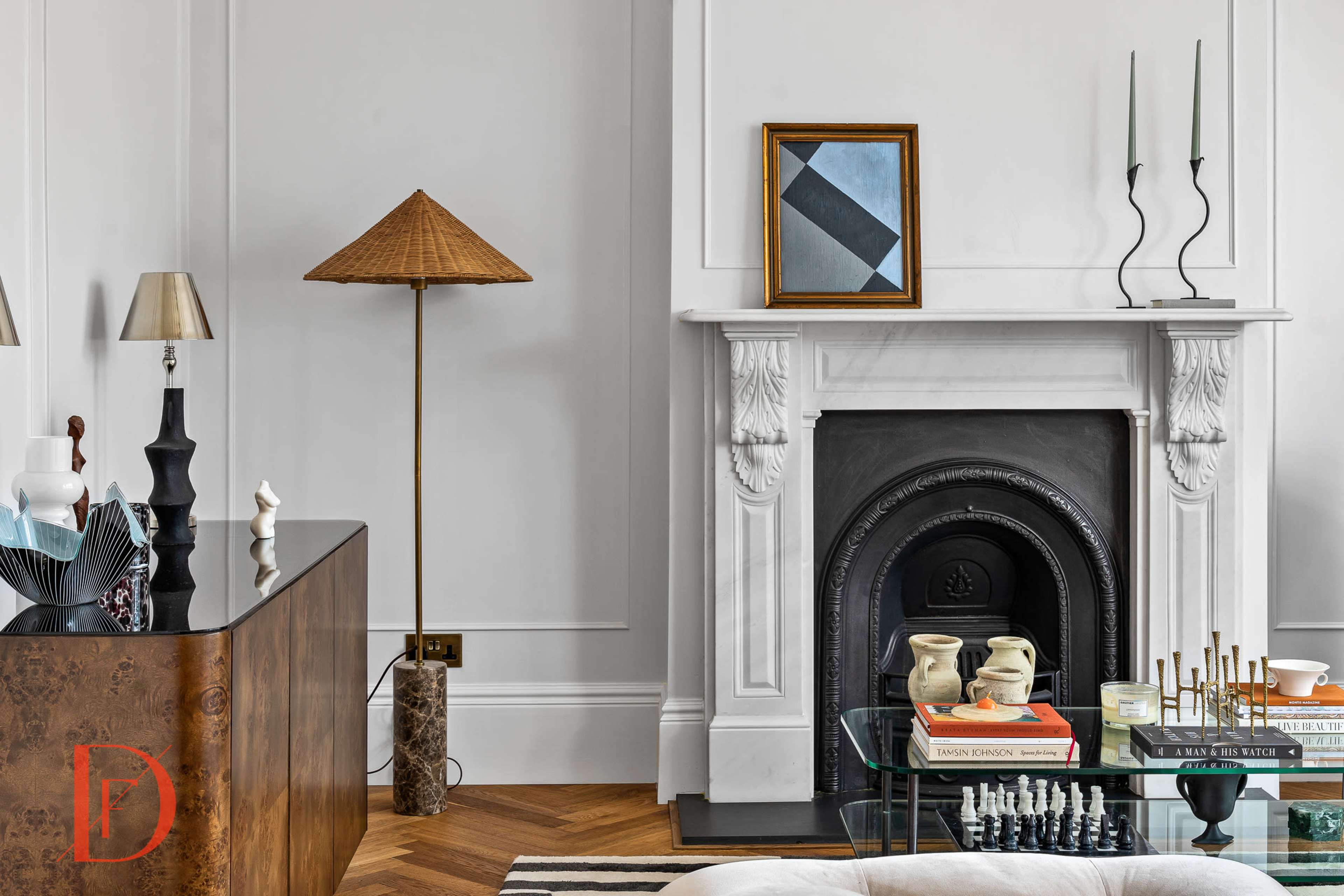 A modern living room featuring a fireplace with a black insert, a glass coffee table stacked with books, and decorative items on a wooden sideboard, all under a neutral-colored wall.