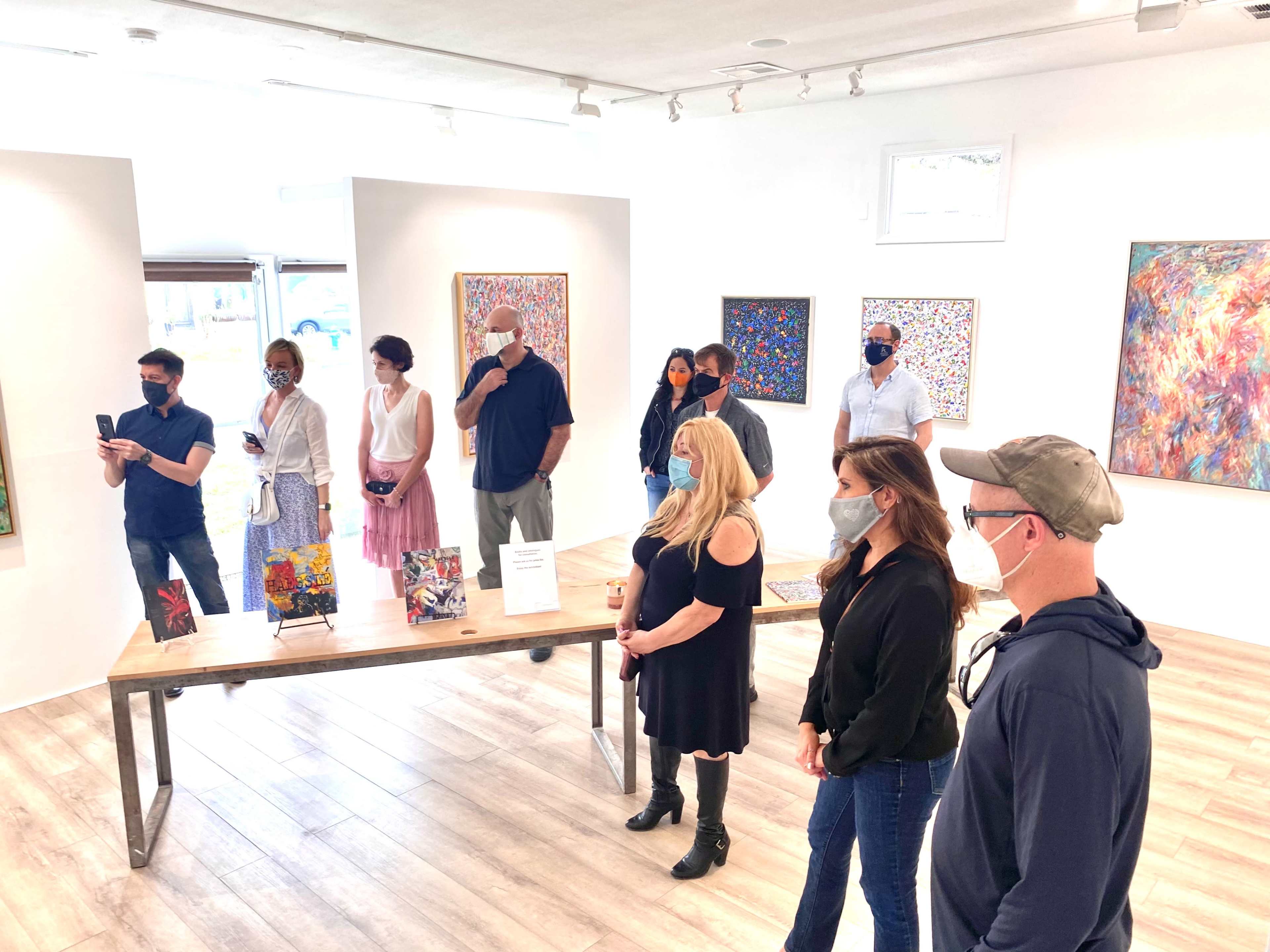 A group of people wearing masks stands in an art gallery, observing various colorful paintings displayed on the walls.