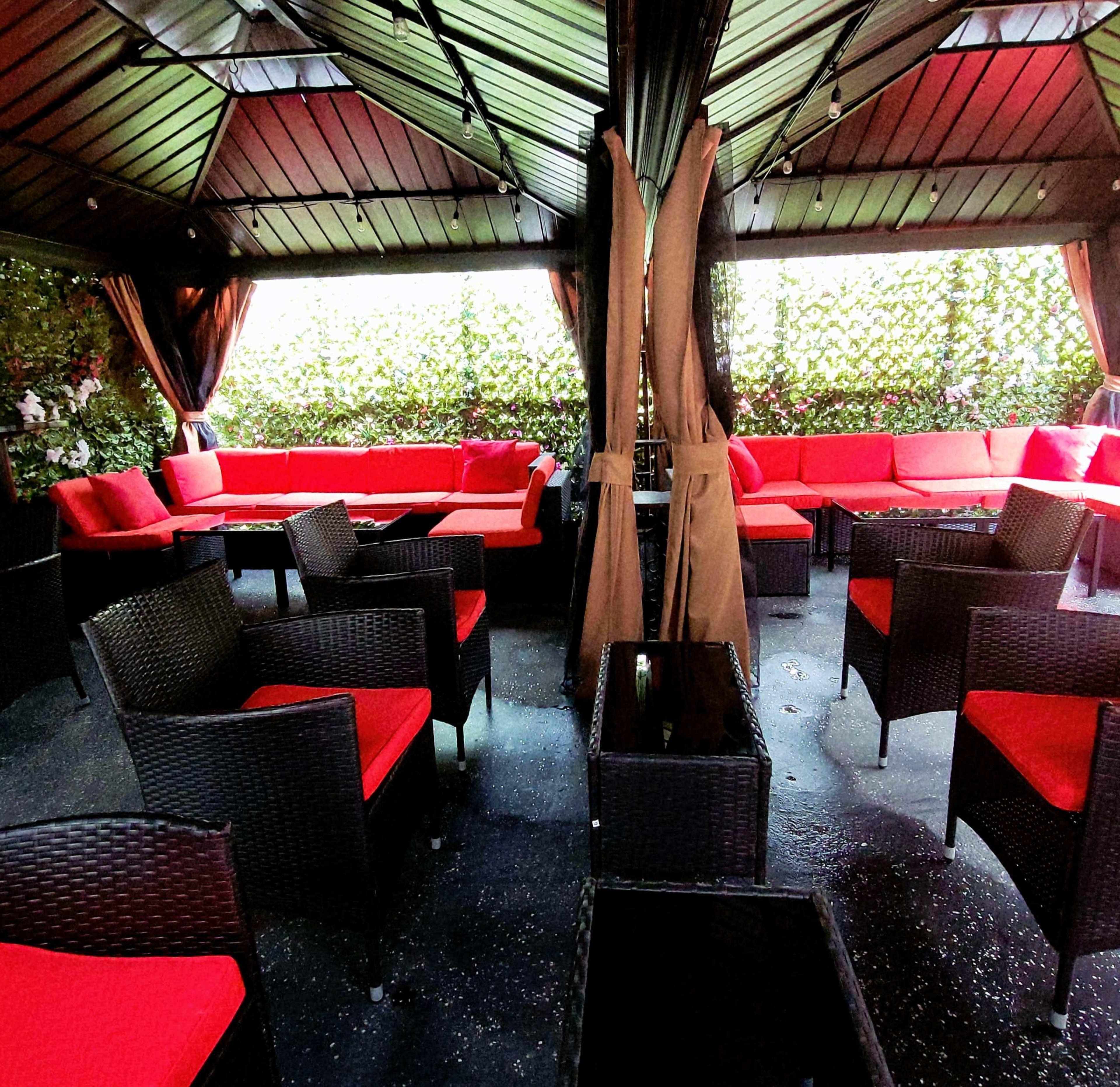 A sheltered outdoor seating area features red cushions on black wicker furniture amidst lush greenery.