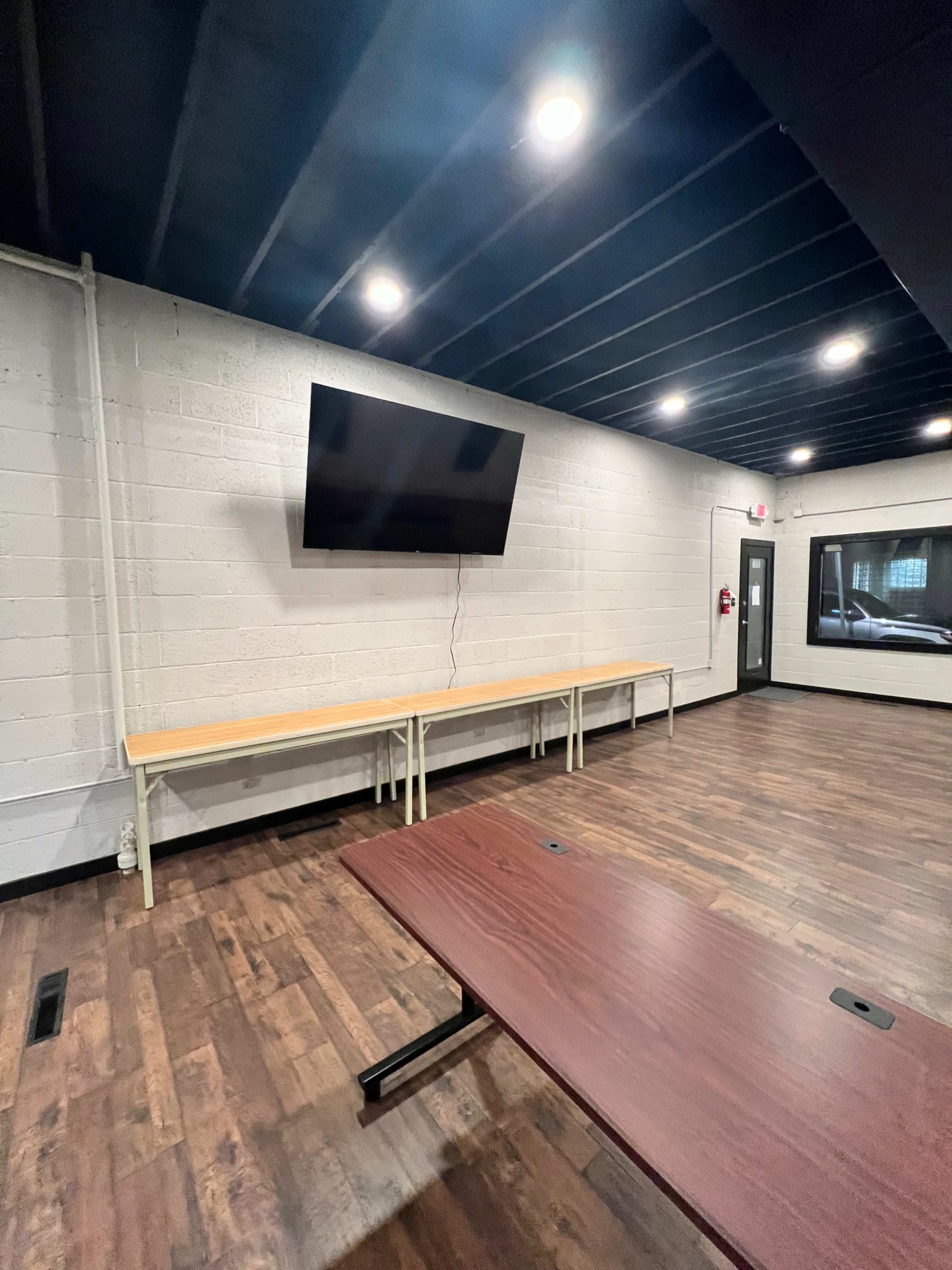 A modern room with wooden flooring, a wall-mounted television, a long table along one wall, and a dark ceiling.