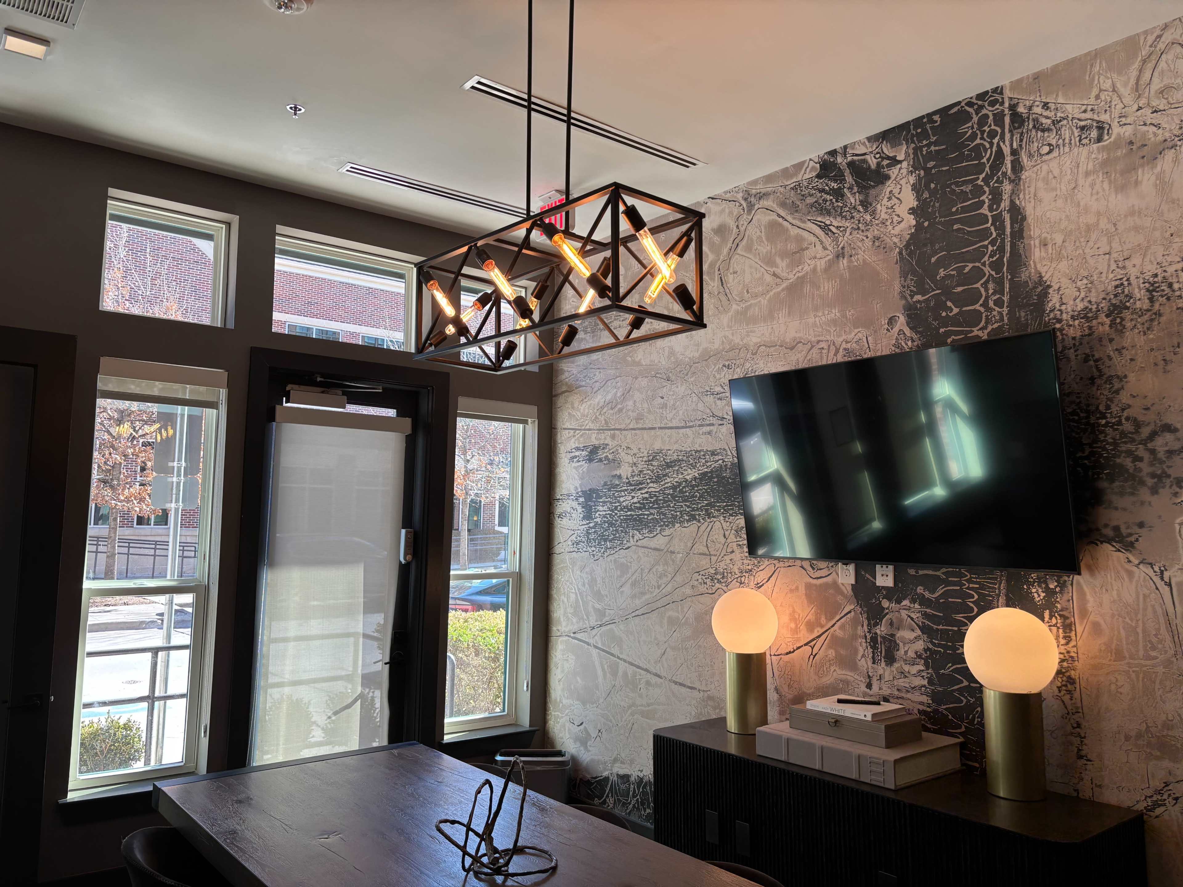 The image shows a modern room with large windows, a wall-mounted TV, a decorative light fixture, and two spherical lamps on a dark wooden side table.