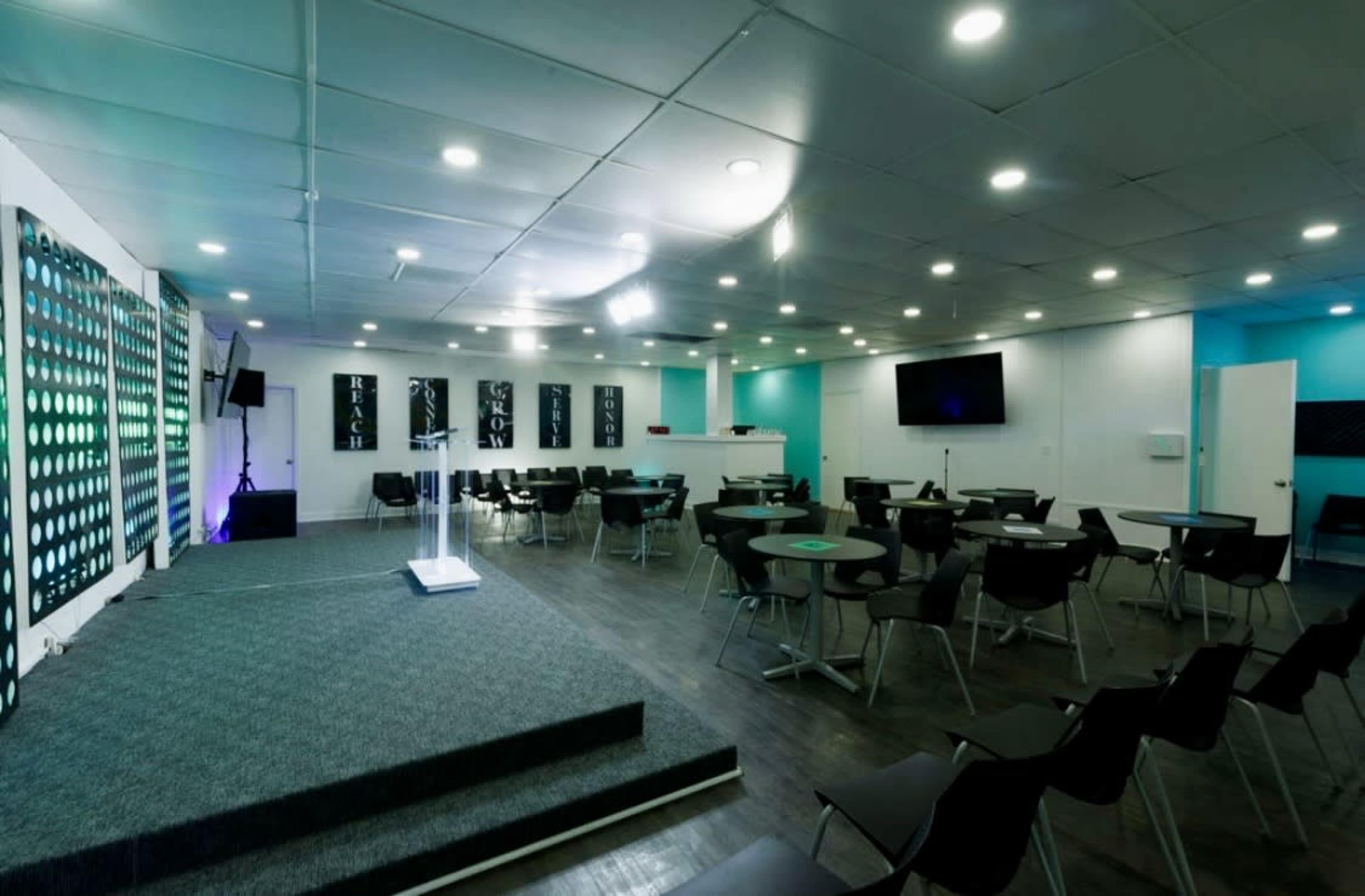 The image shows a modern event space with a stage, rows of black chairs, and a bar area in the background.