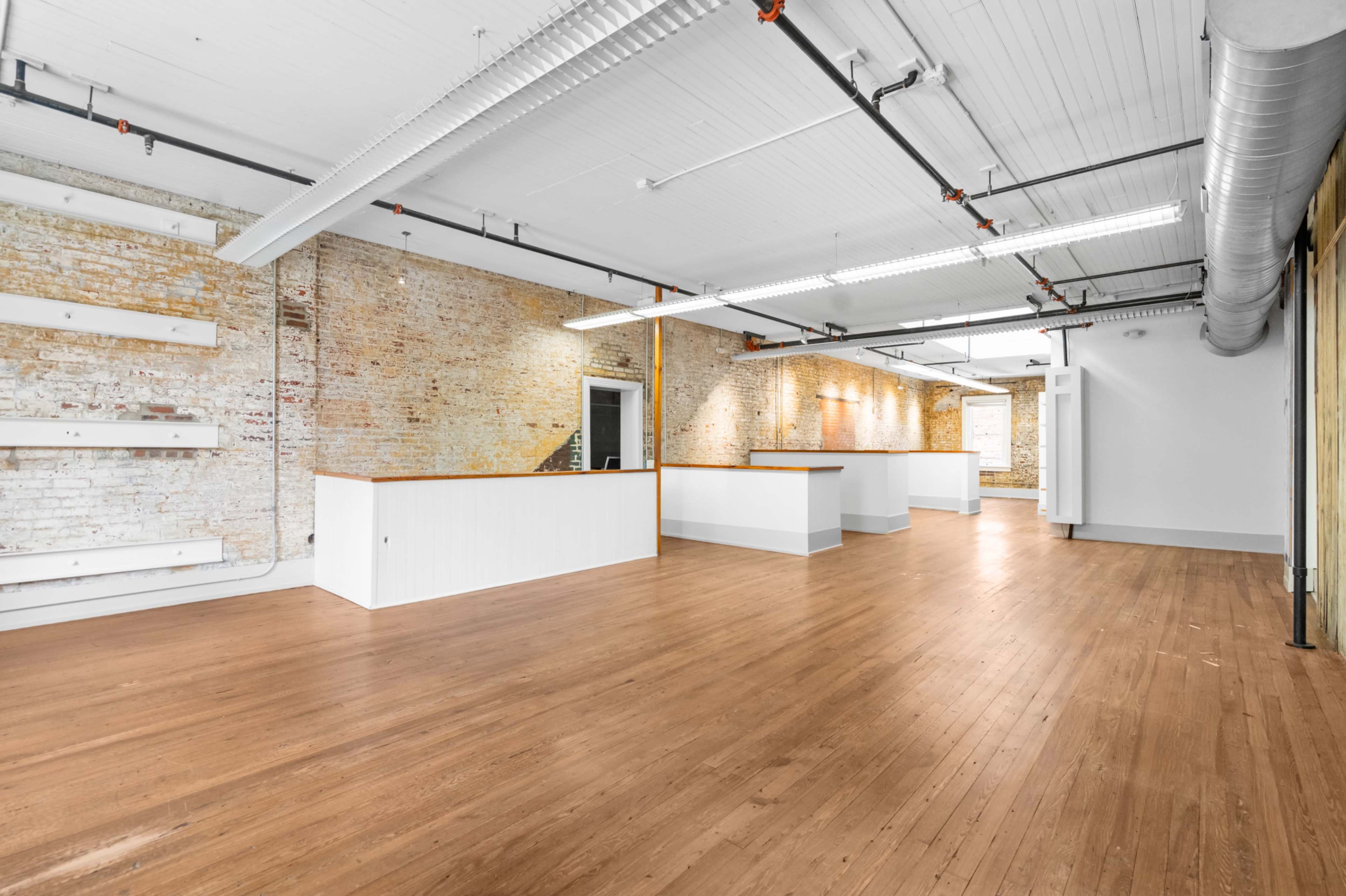 The image shows an empty, spacious interior of a commercial space with white walls, wooden flooring, and exposed brick accents.