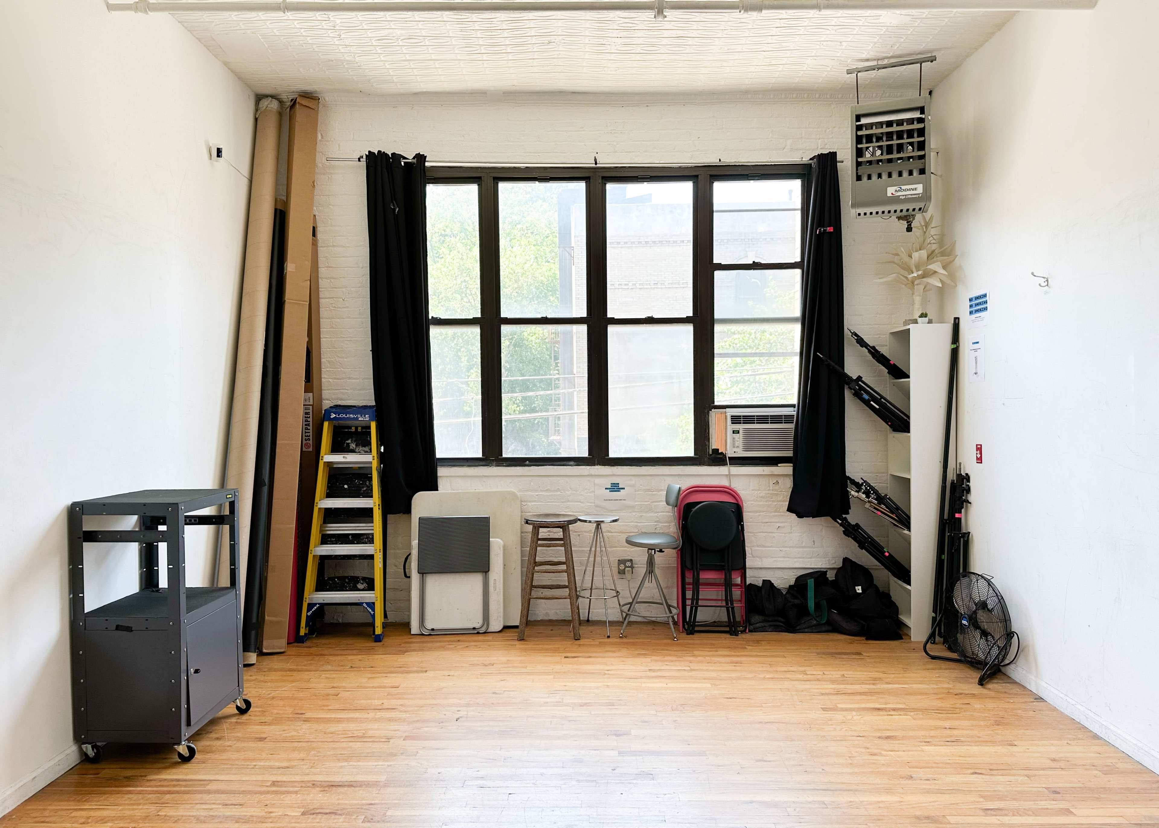 An empty room with wooden floors, featuring a large window with black curtains, a ladder, various stacked items, and a small cart in the corner.