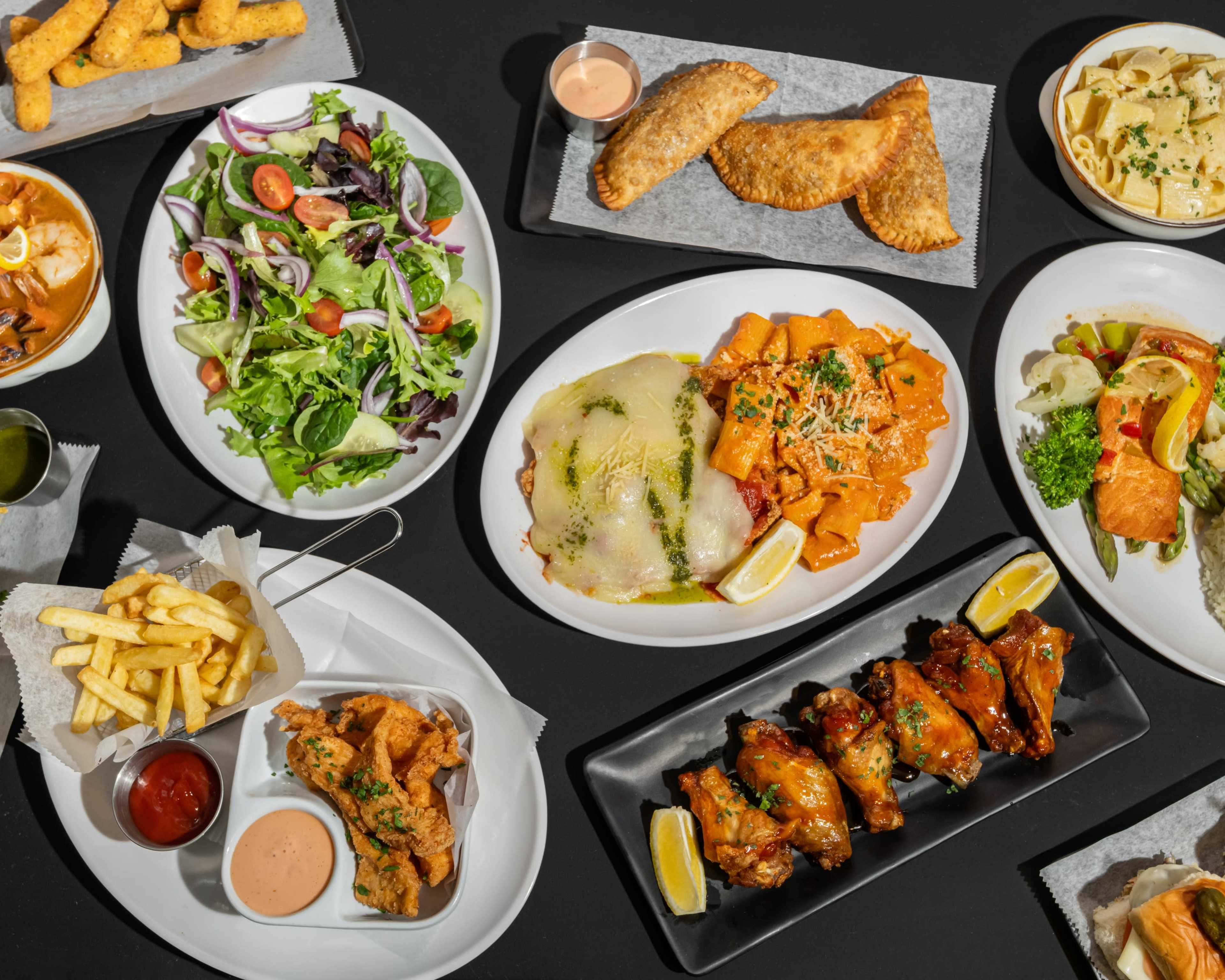 The image displays a variety of dishes arranged on a table, including salads, pasta, chicken wings, and fried items.