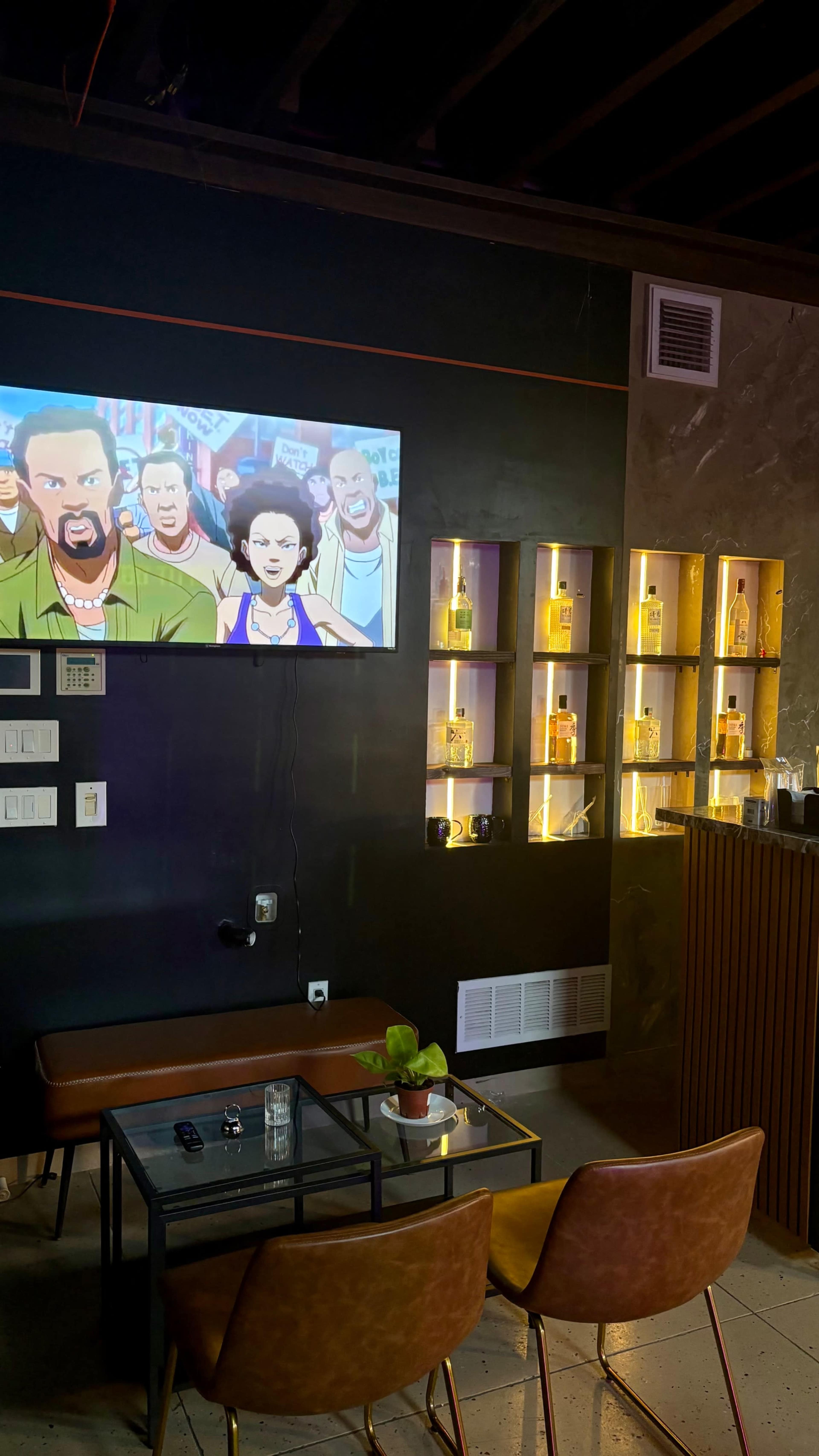 The image shows a wall featuring a large television displaying an animated scene, with a cozy seating area in front that includes a glass table and two chairs, along with illuminated shelves displaying bottles.