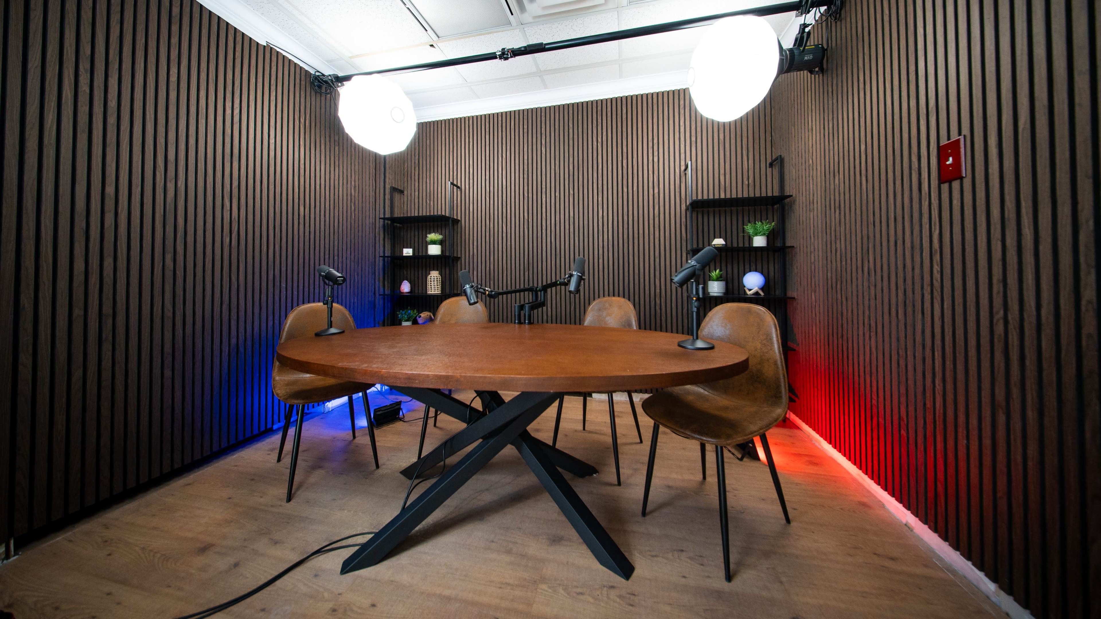 A well-lit podcast recording studio features a round wooden table surrounded by five chairs, with microphones positioned at each seat and LED lights casting a glow on the walls.