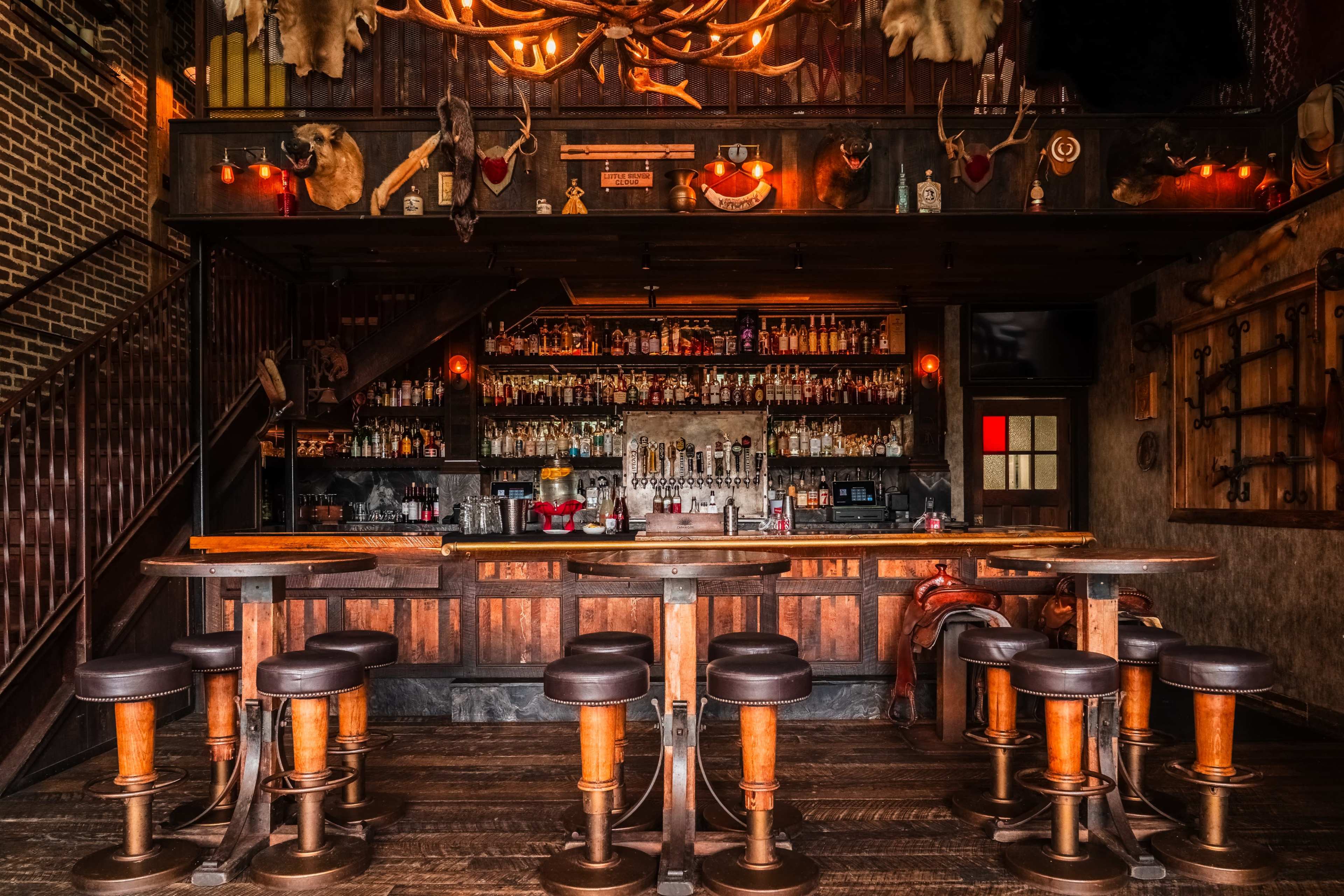 The image shows a rustic bar with wooden stools arranged in front of a well-stocked shelf of liquor bottles and antique decorations on the walls.