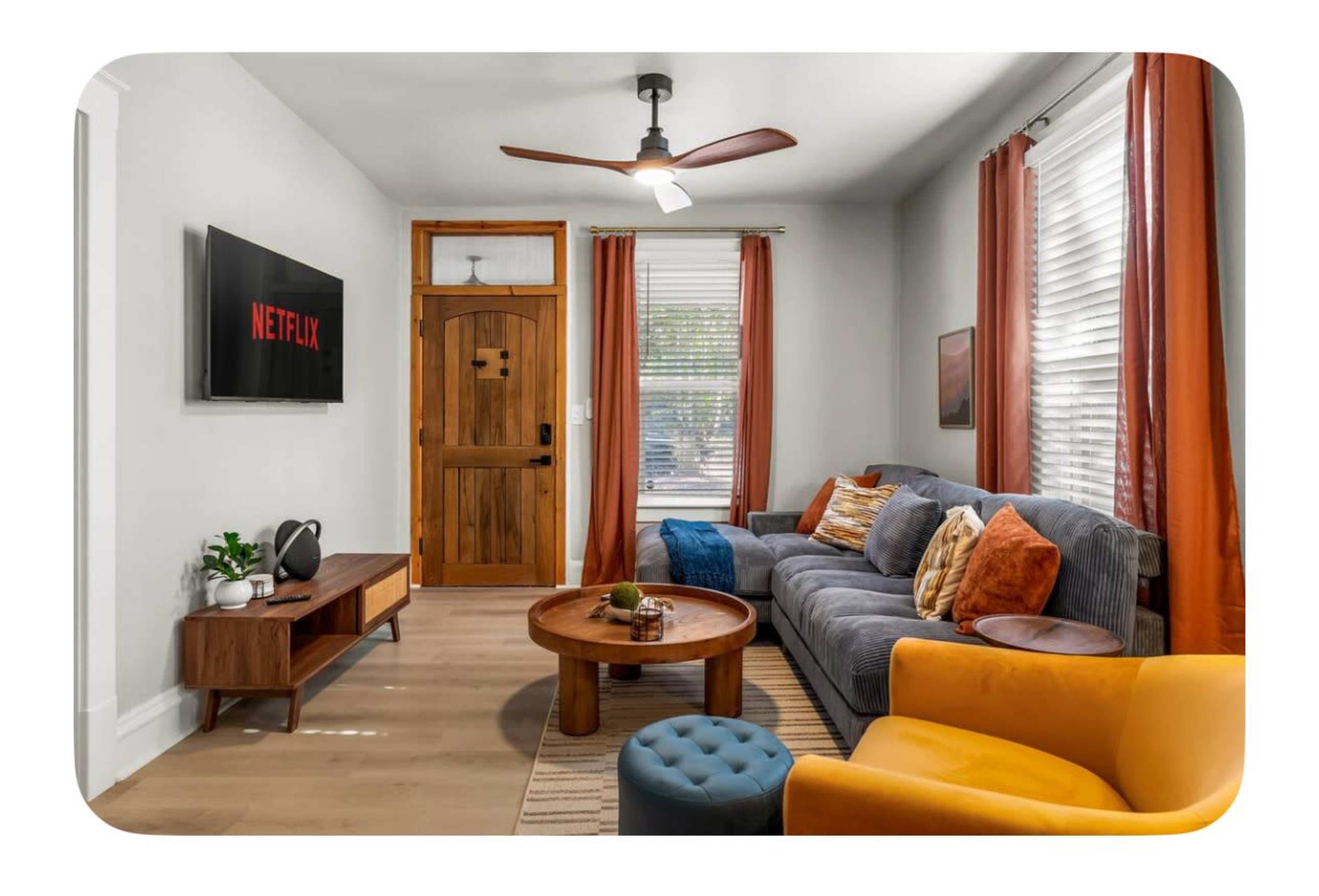 A cozy living room features a gray sofa, an orange armchair, a wooden coffee table, and a wall-mounted TV displaying the Netflix logo, with natural light coming through the windows.
