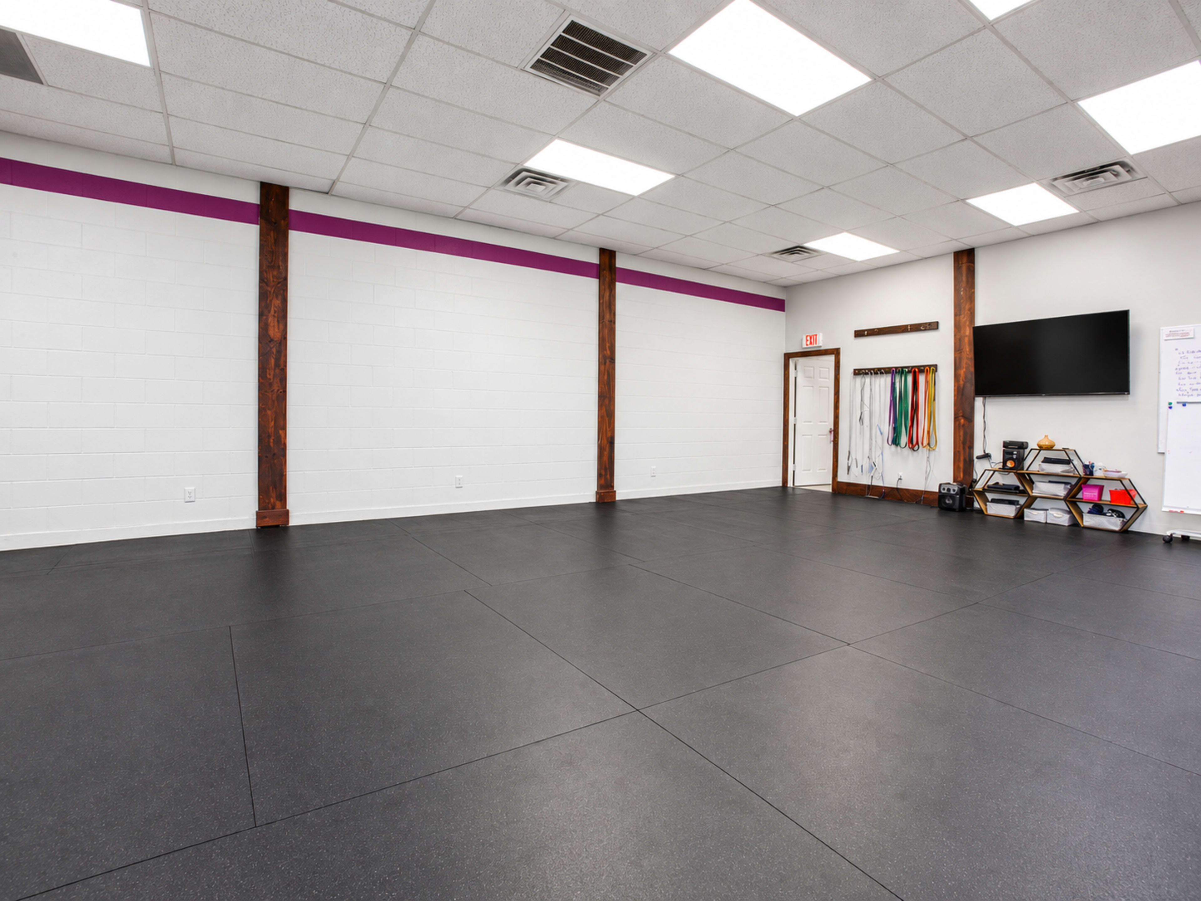The image shows an empty fitness studio with black rubber flooring, white walls, and a few exercise accessories on shelves.