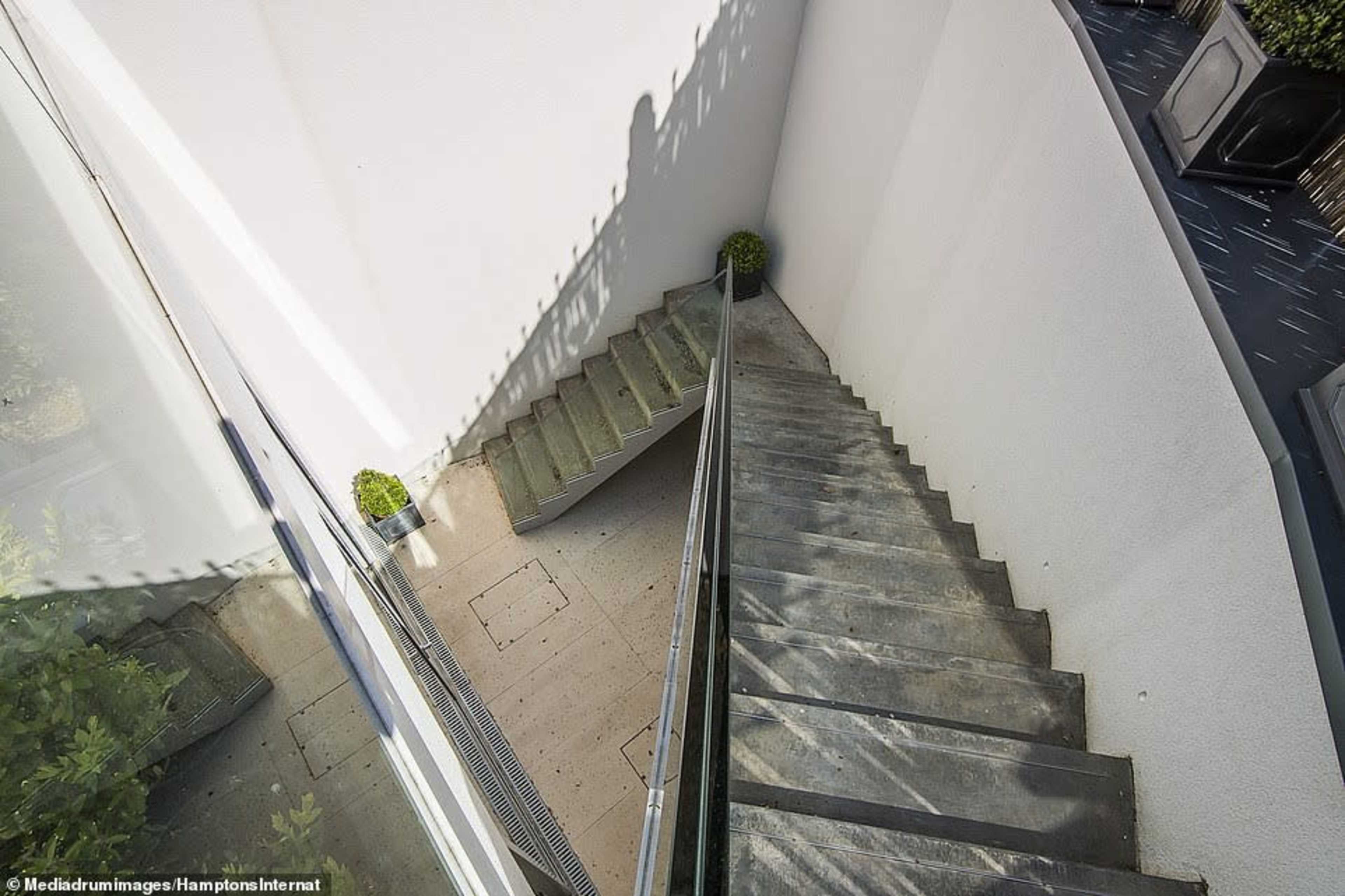 A concrete staircase leading down in a minimalist outdoor setting, bordered by a glass railing and a white wall.