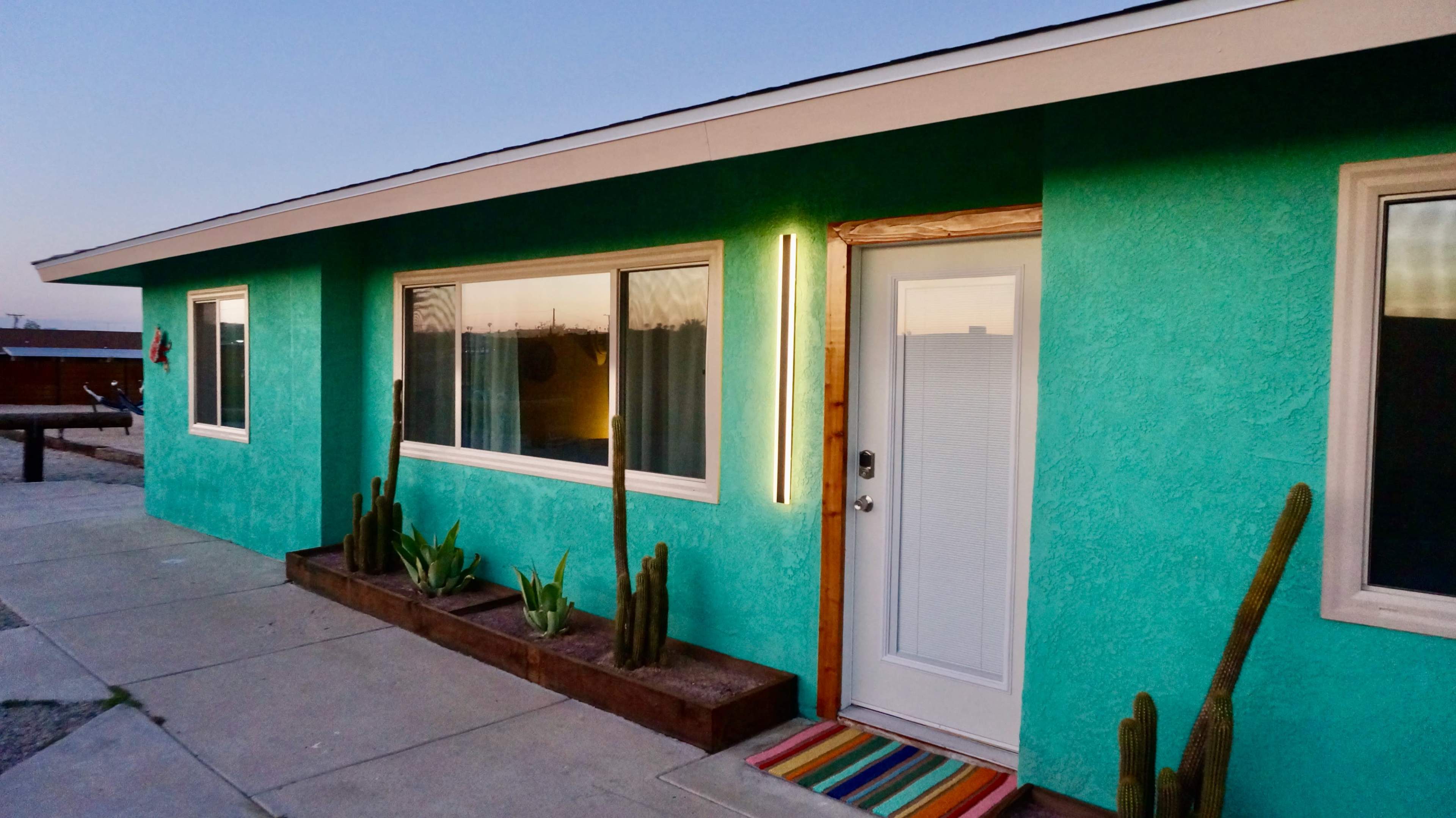 A turquoise-colored house with a modern door and cacti planted in a raised bed by the entrance.