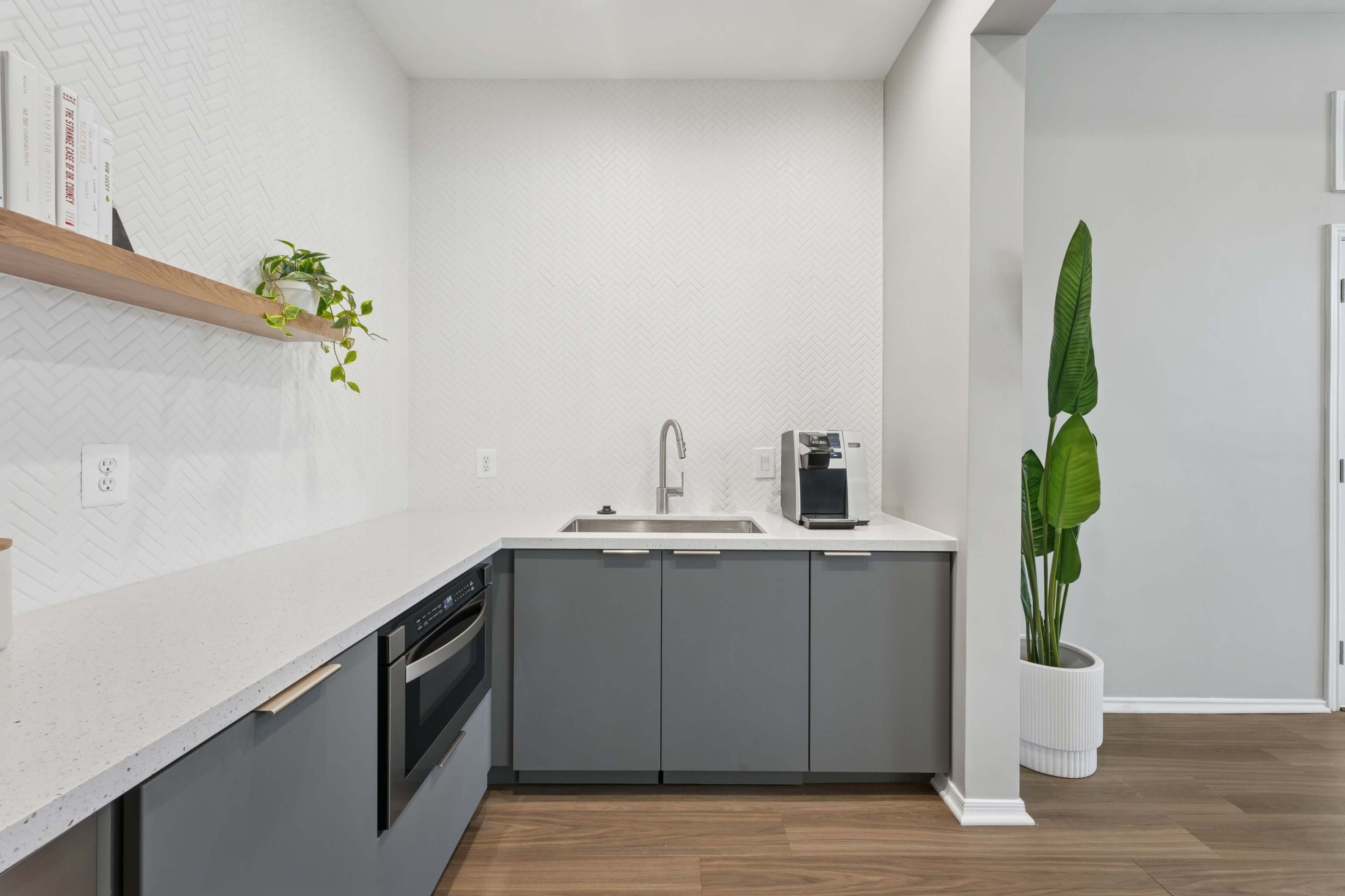 A modern kitchen features gray cabinets, a white countertop, a sink, and a coffee maker, with a plant on a shelf and a tall potted plant in one corner.