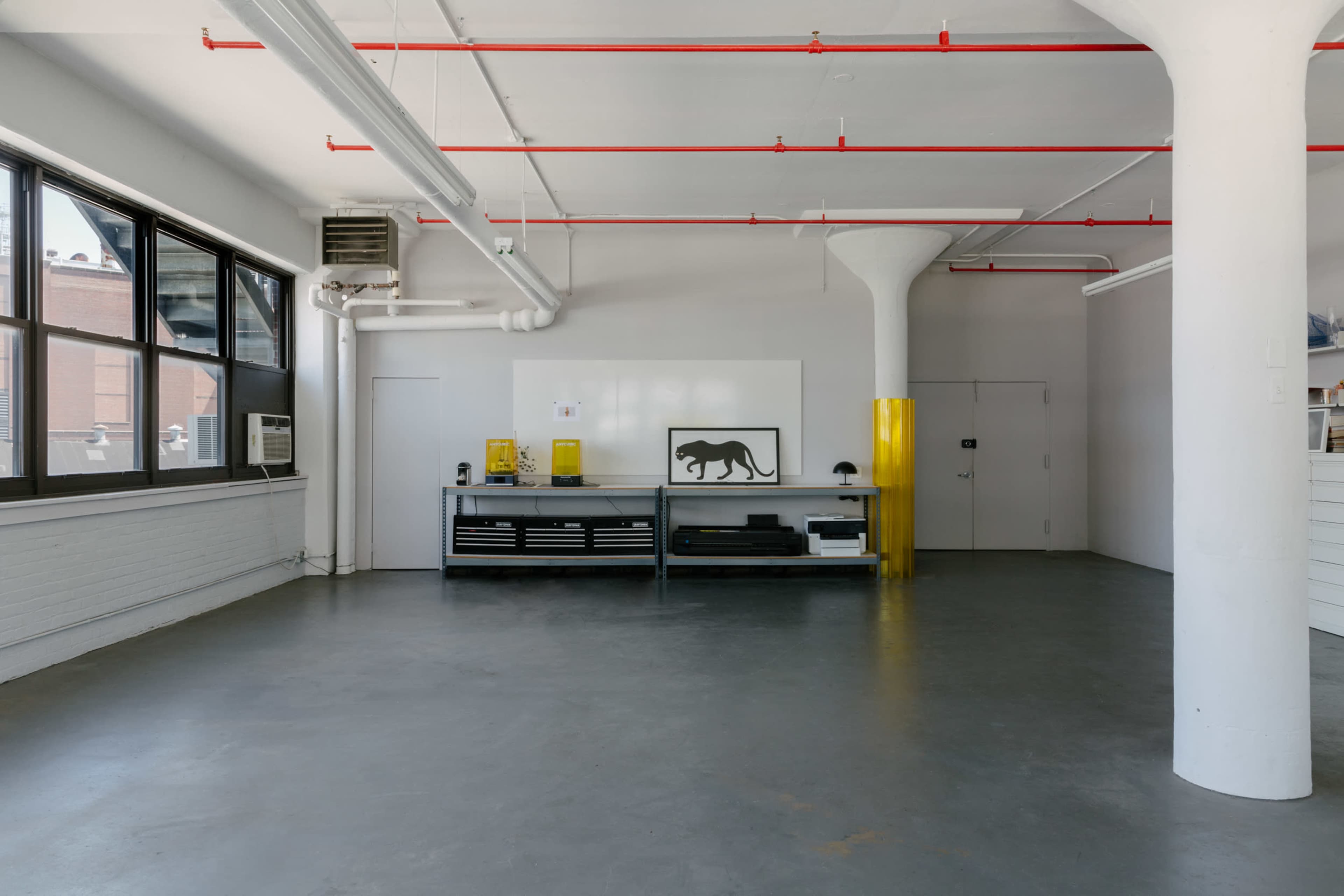 An industrial-style studio space with large windows, exposed piping, a central work table, and minimalist decor.
