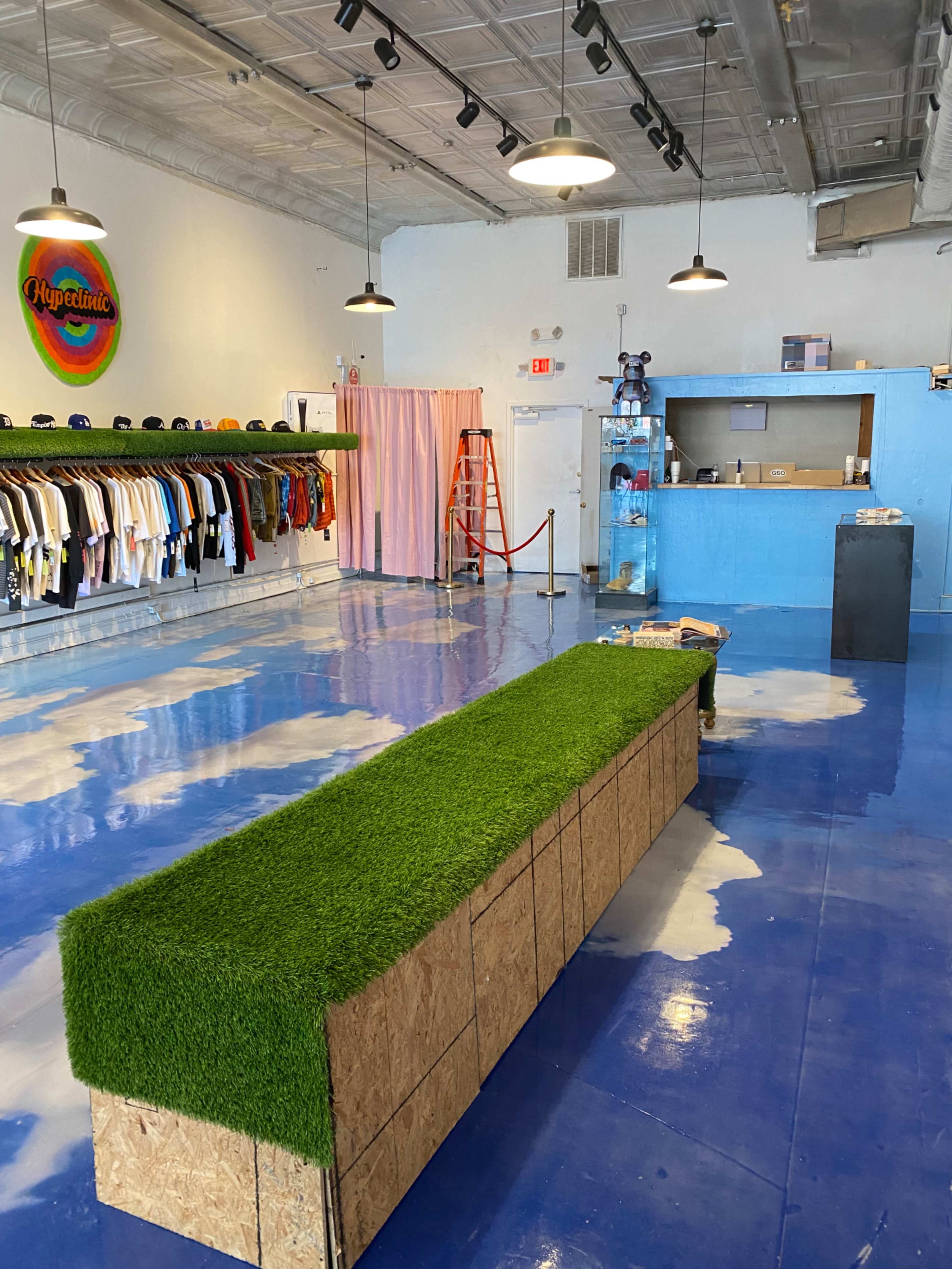 A retail space with clothing displayed on racks, a long green grass-like bench in the center, and a blue counter at the back.
