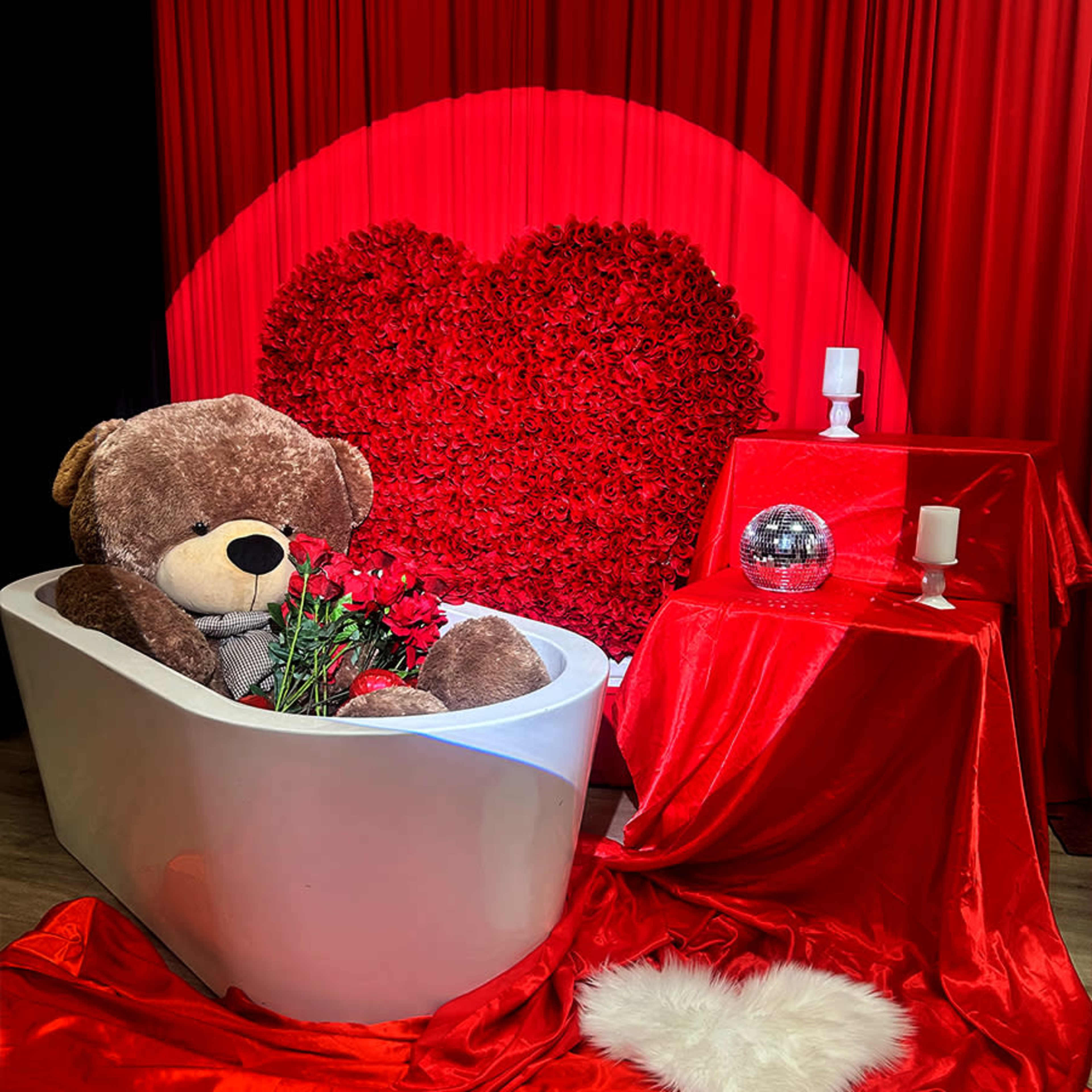 A large teddy bear sits in a white bathtub surrounded by red roses, with a heart-shaped backdrop and decorative red drapes.