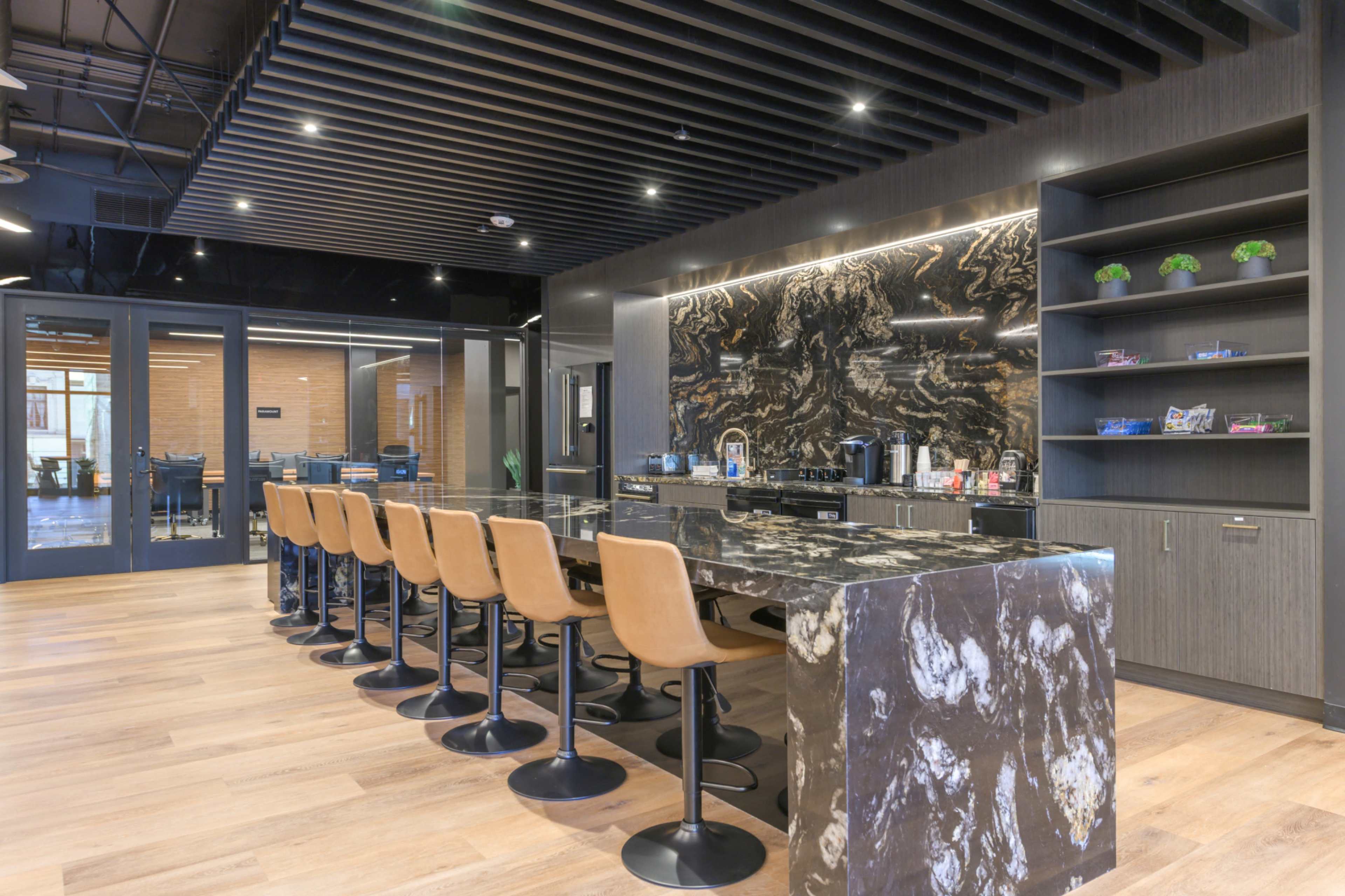 The image shows a modern kitchen area with a large marble countertop, several high-backed stools, and a dark wooden ceiling.